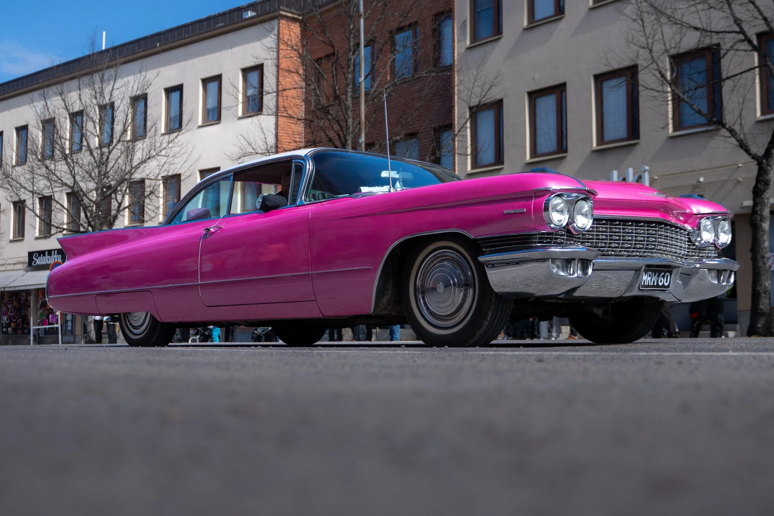 A vintage pink Cadillac car parked on the street, with a low-angle view showing the front and side of the vehicle, against a cityscape background.