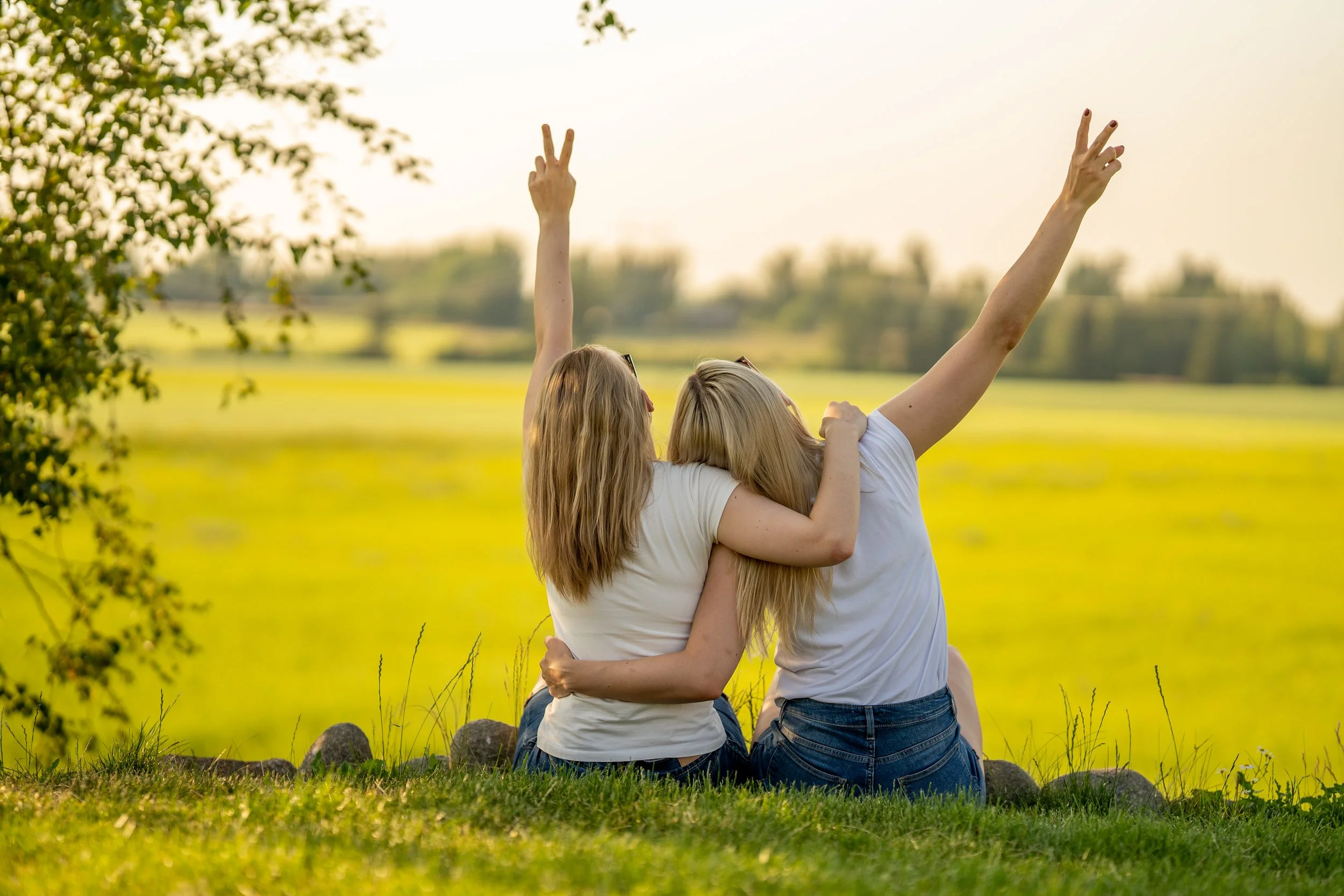 Two women with blonde hair sitting on grass, facing away, hugging and making peace signs with their hands, in a field with yellow flowers.