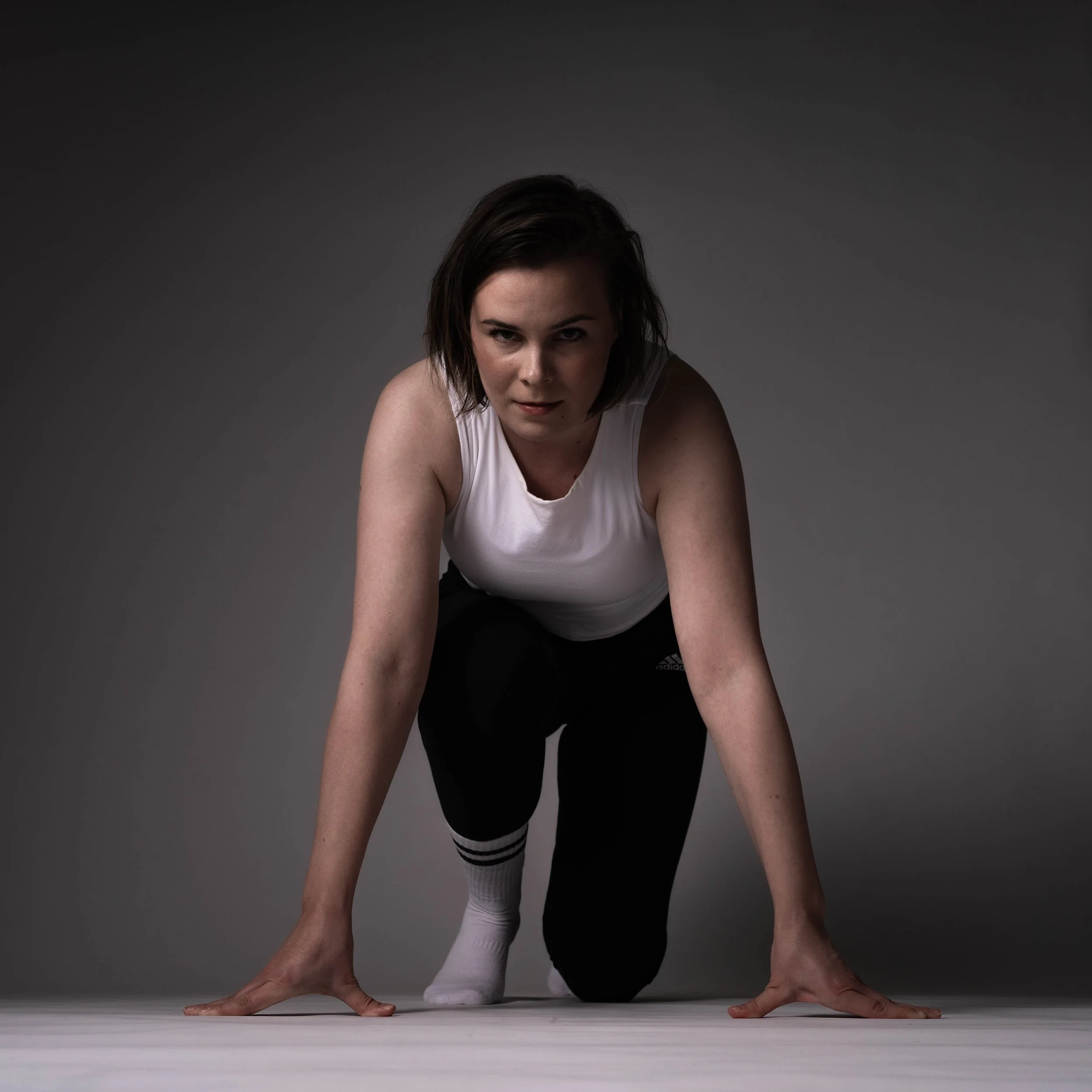 Young woman in athletic clothing crouched in starting position on a plain gray background.