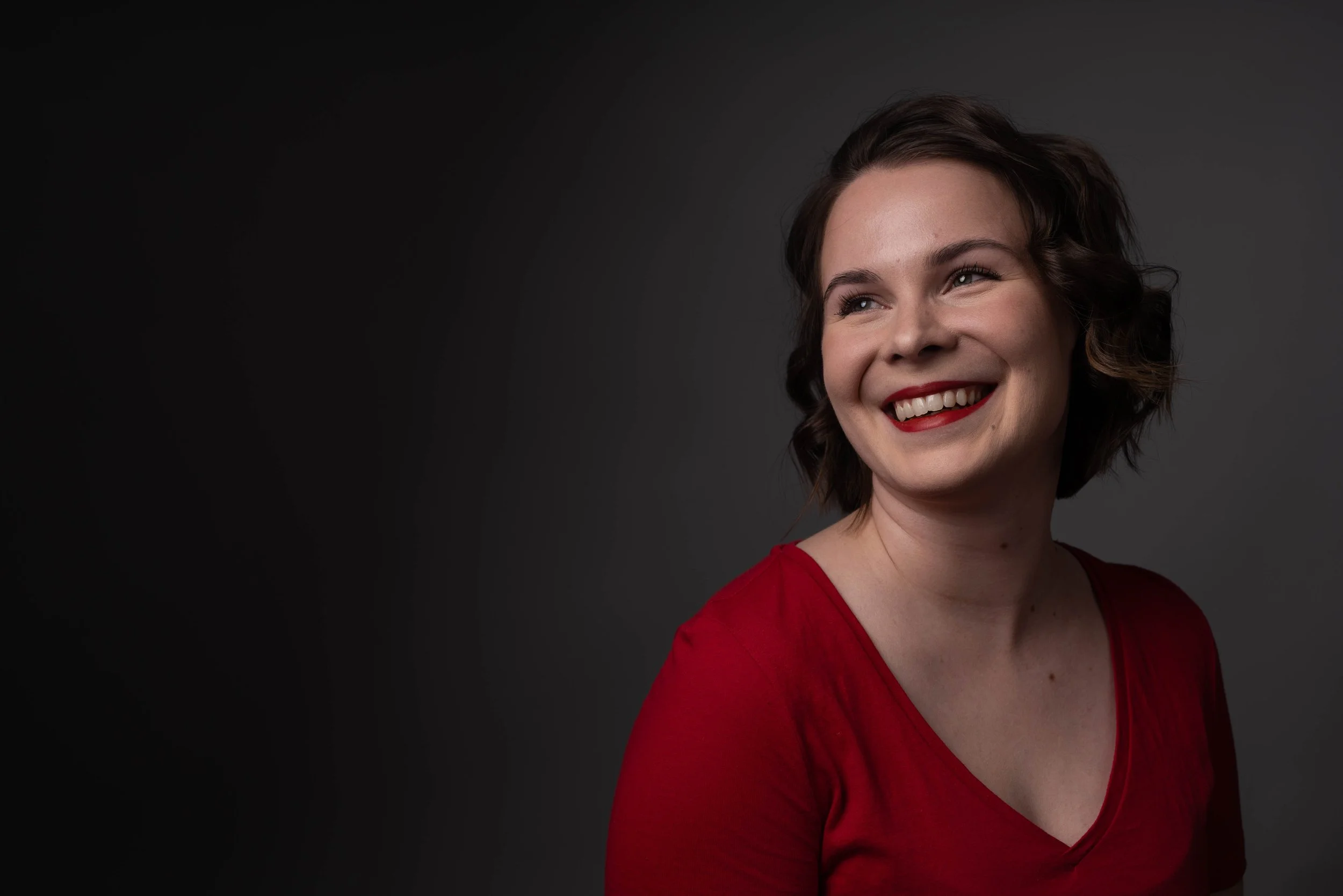A smiling woman with short brown hair wearing a red top against a dark gray background.