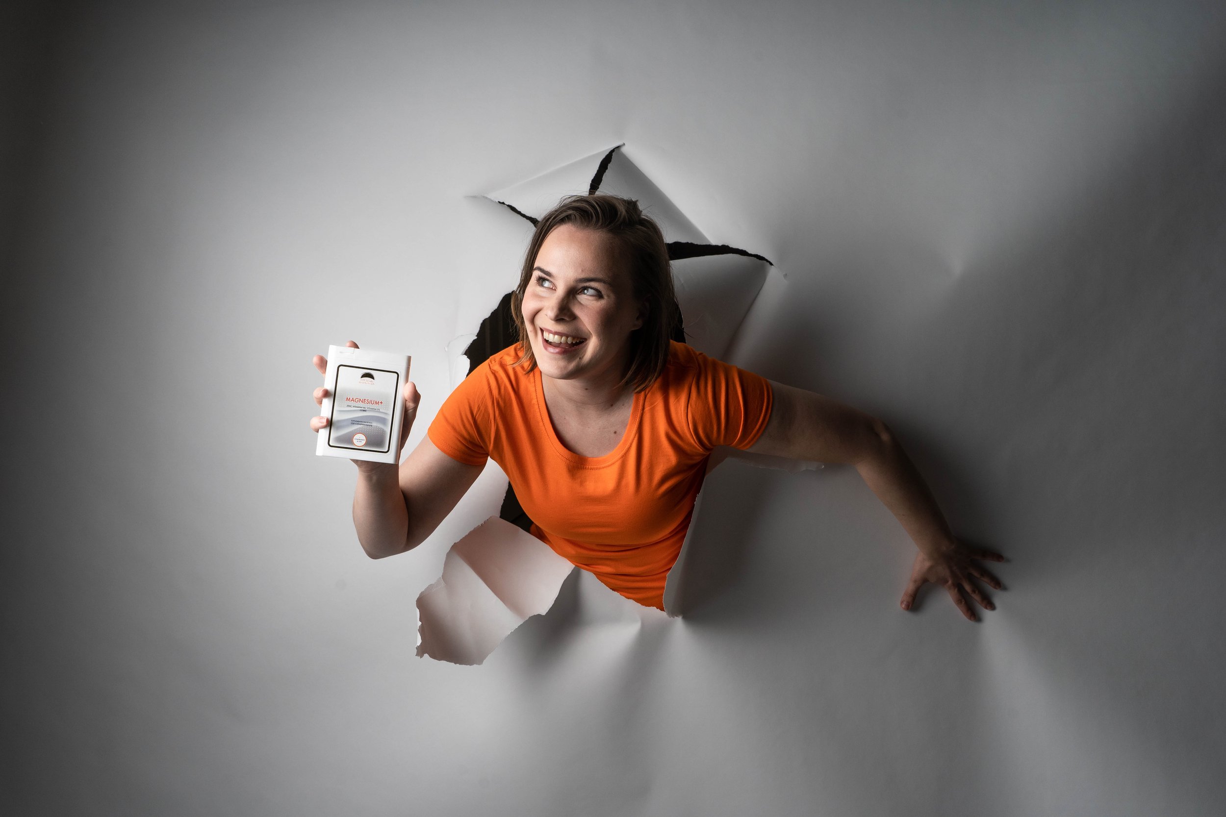 Woman in an orange shirt emerging through torn white paper wall, smiling and holding a box of magnesium supplements.