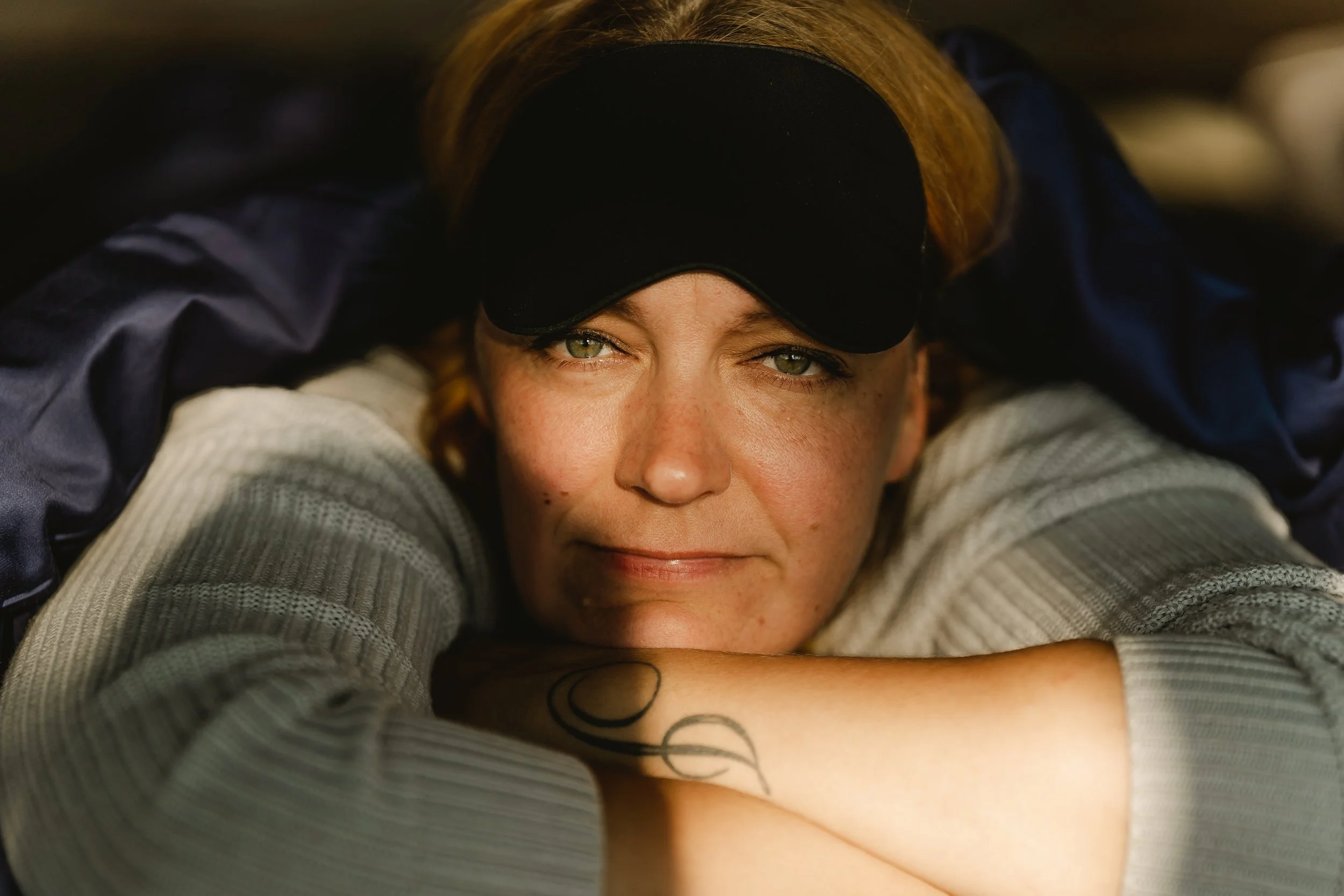 Close-up of a woman with green eyes, wearing a black eye mask pushed up on her forehead, lying down with her arms crossed and resting her chin on them. She has a tattoo of intersecting circles on her forearm, wearing a light gray, ribbed long sleeve 