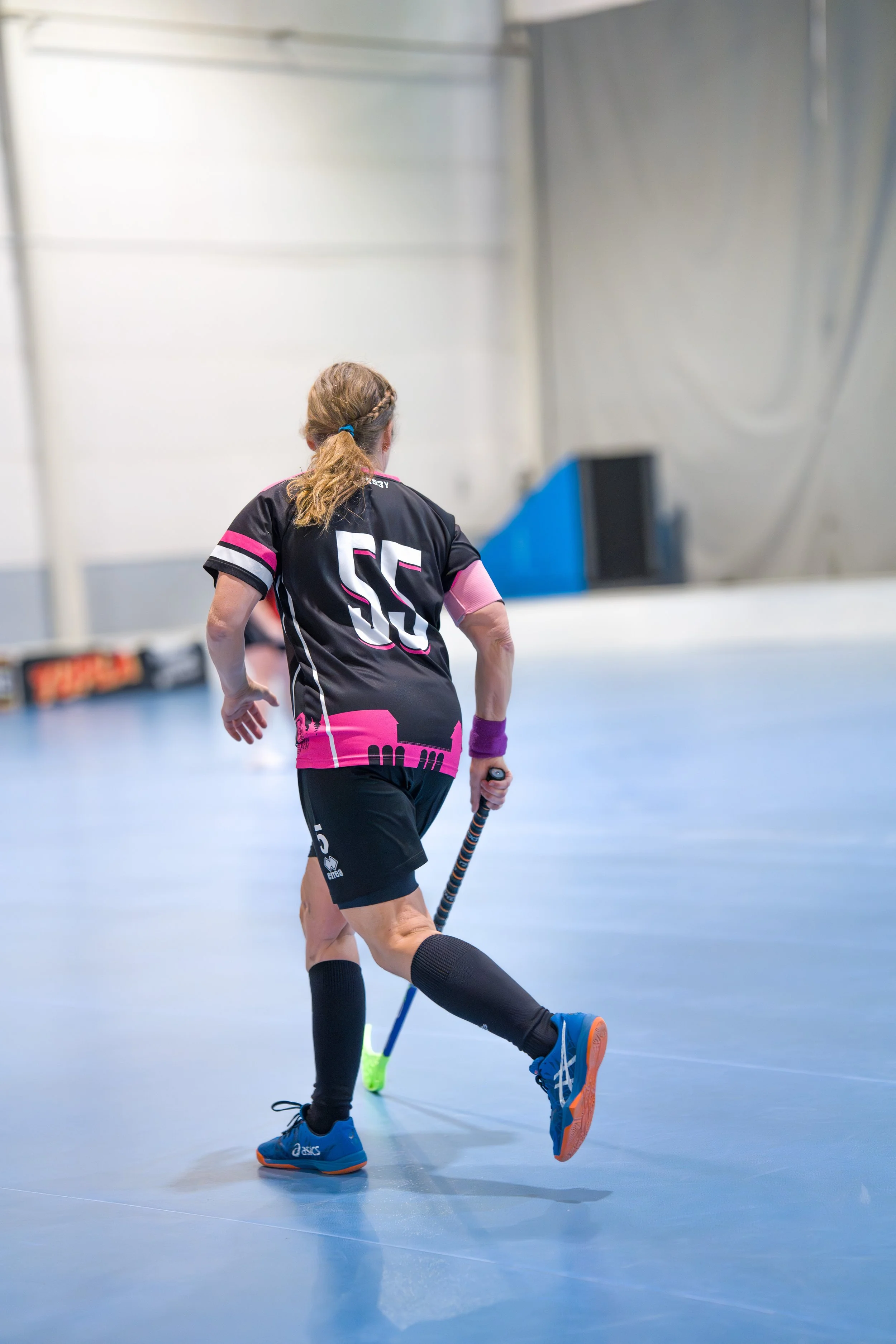 A female floorball player with brown hair in a ponytail, wearing a black and pink uniform, holding a floorball stick, playing indoors on a light blue court.