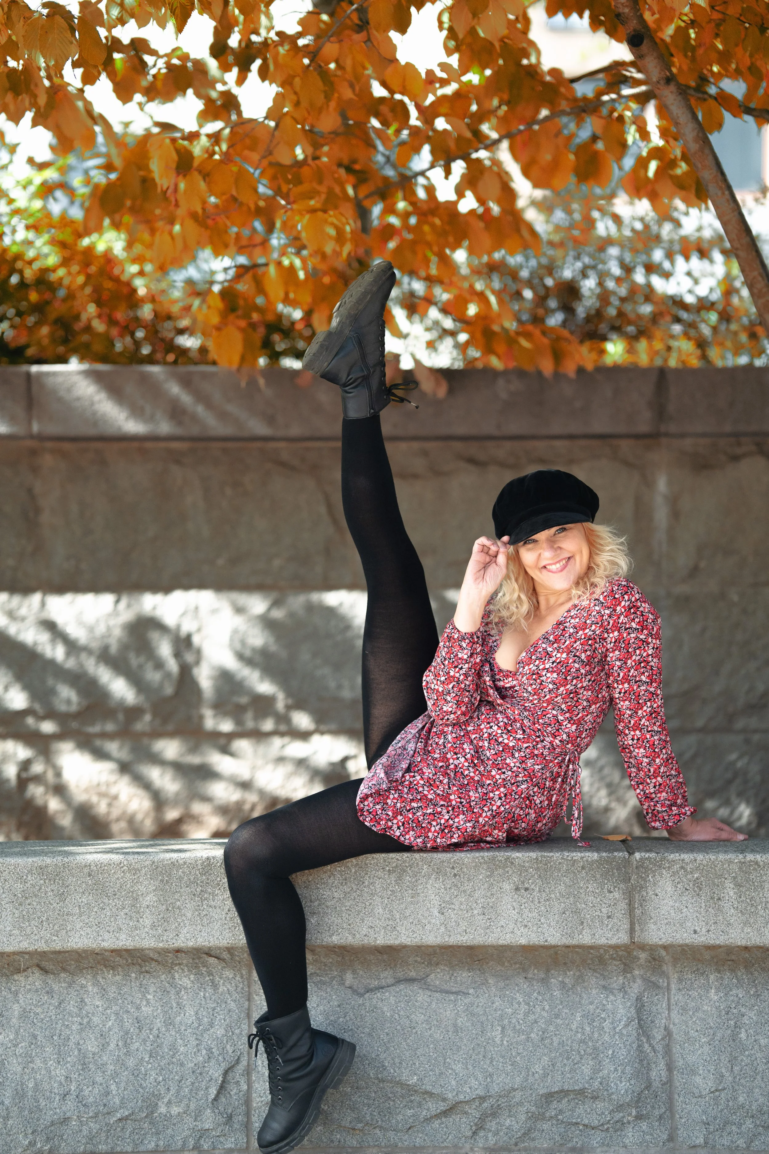 A woman with blonde curly hair, wearing a black hat, red floral dress, black tights, and black boots, sitting on a stone ledge with one leg raised straight up and smiling at the camera outdoors, with autumn-colored leaves overhead.