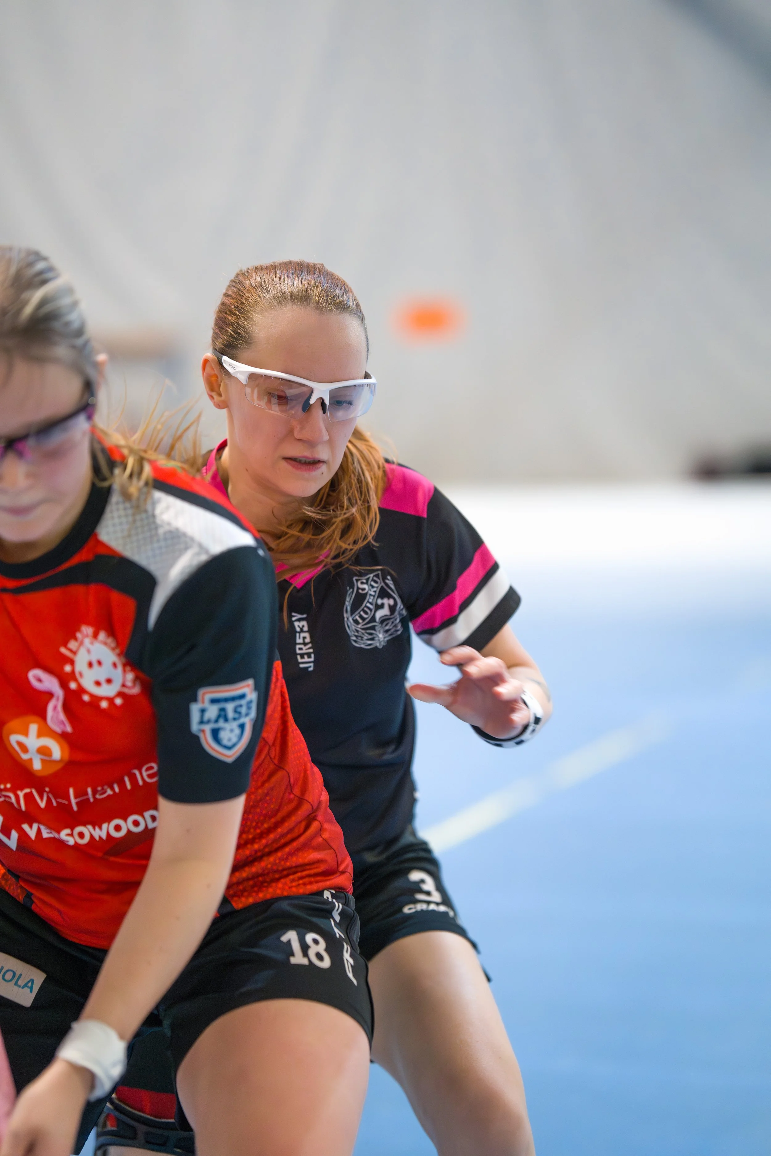 Two female wheelchair rugby players in action during a game indoors. One wearing a red jersey and the other in a black jersey, both wearing protective goggles.