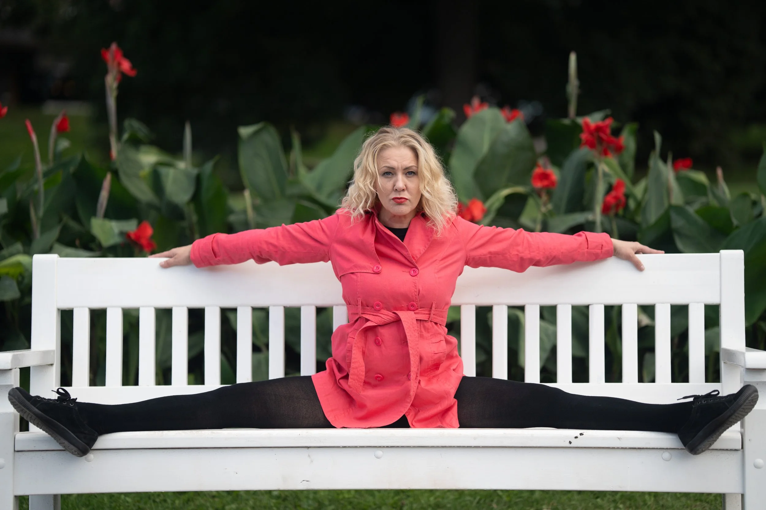 A woman with blonde curly hair wearing a pink trench coat sitting on a white park bench with her legs spread wide apart and arms stretched out, in front of green plants with red flowers.