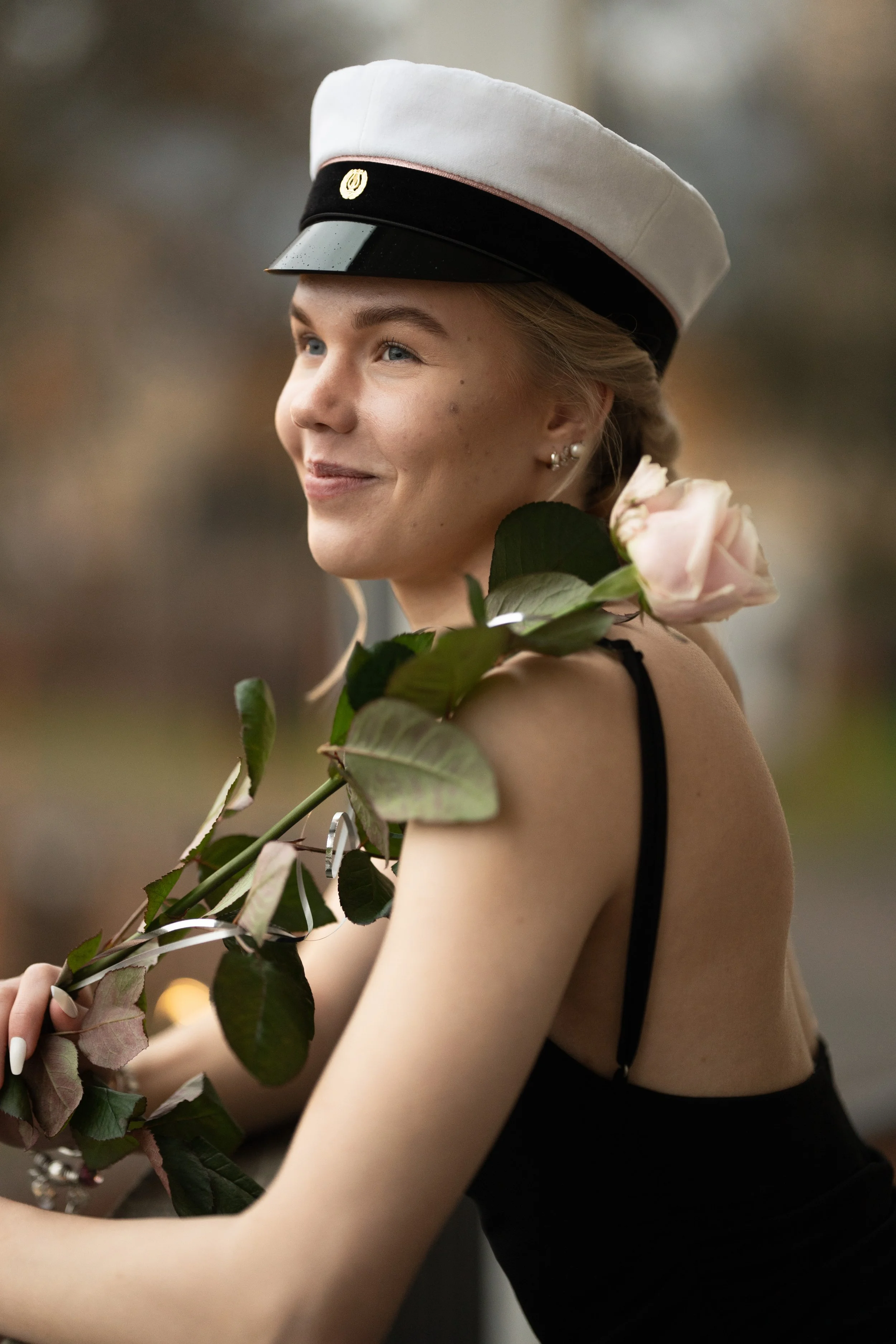 A young woman in a graduation cap holding a pink rose, smiling and looking to the side, outdoors.