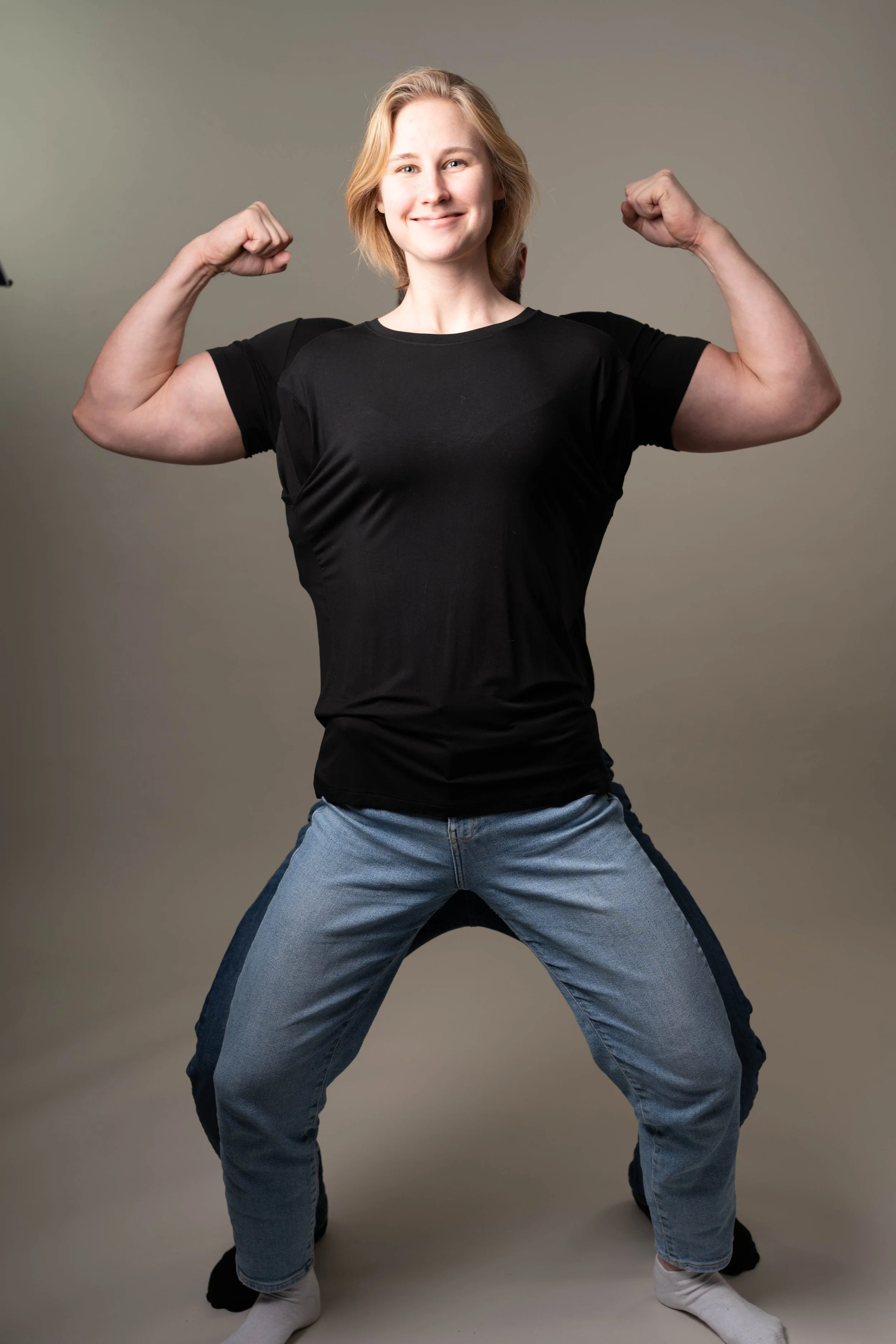 A woman with blonde hair wearing a black T-shirt and jeans is flexing her arms to show her muscles, smiling confidently.