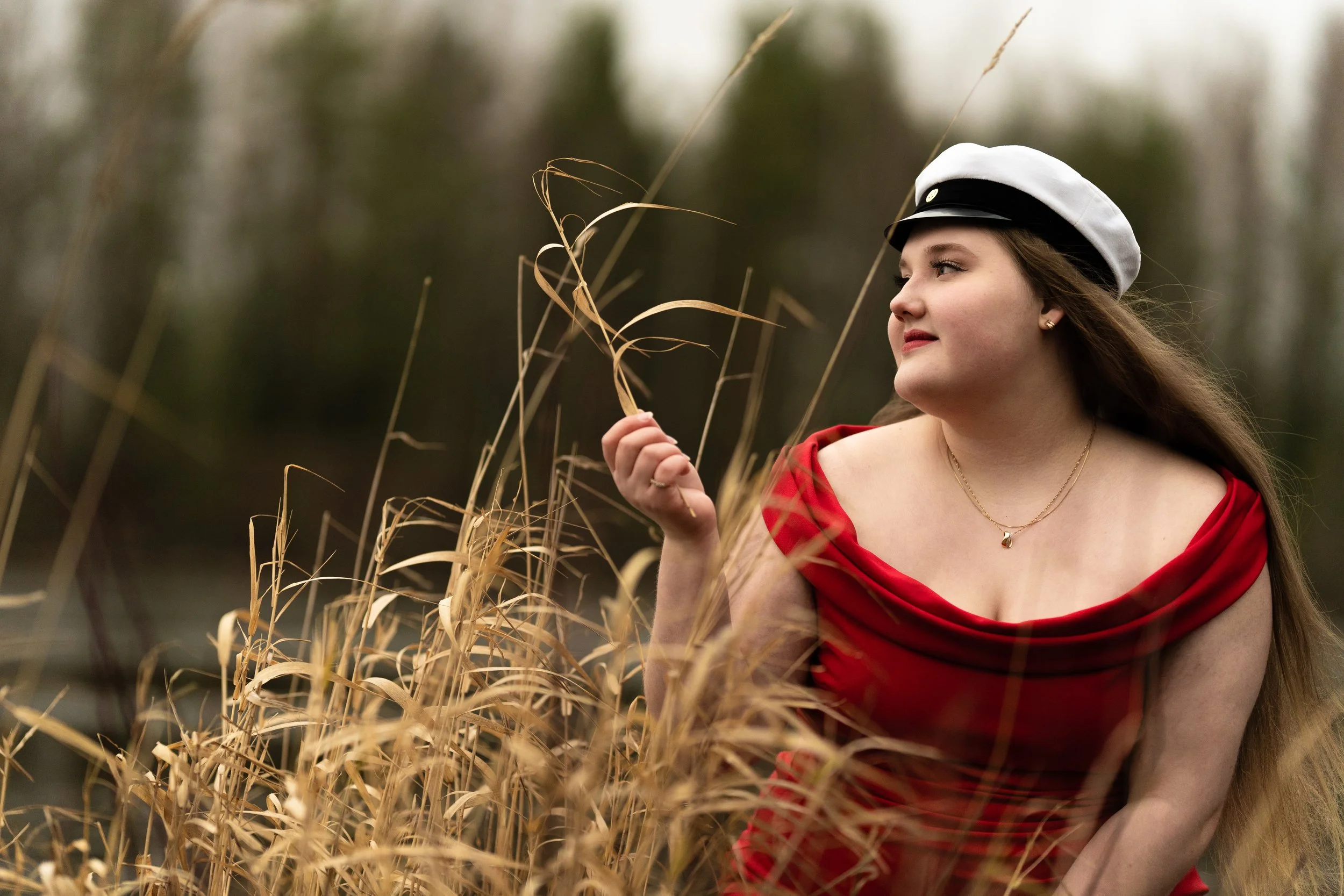 A woman wearing a white cap and a red dress is standing in a field of tall, dry grass, looking to the side with a serene expression.