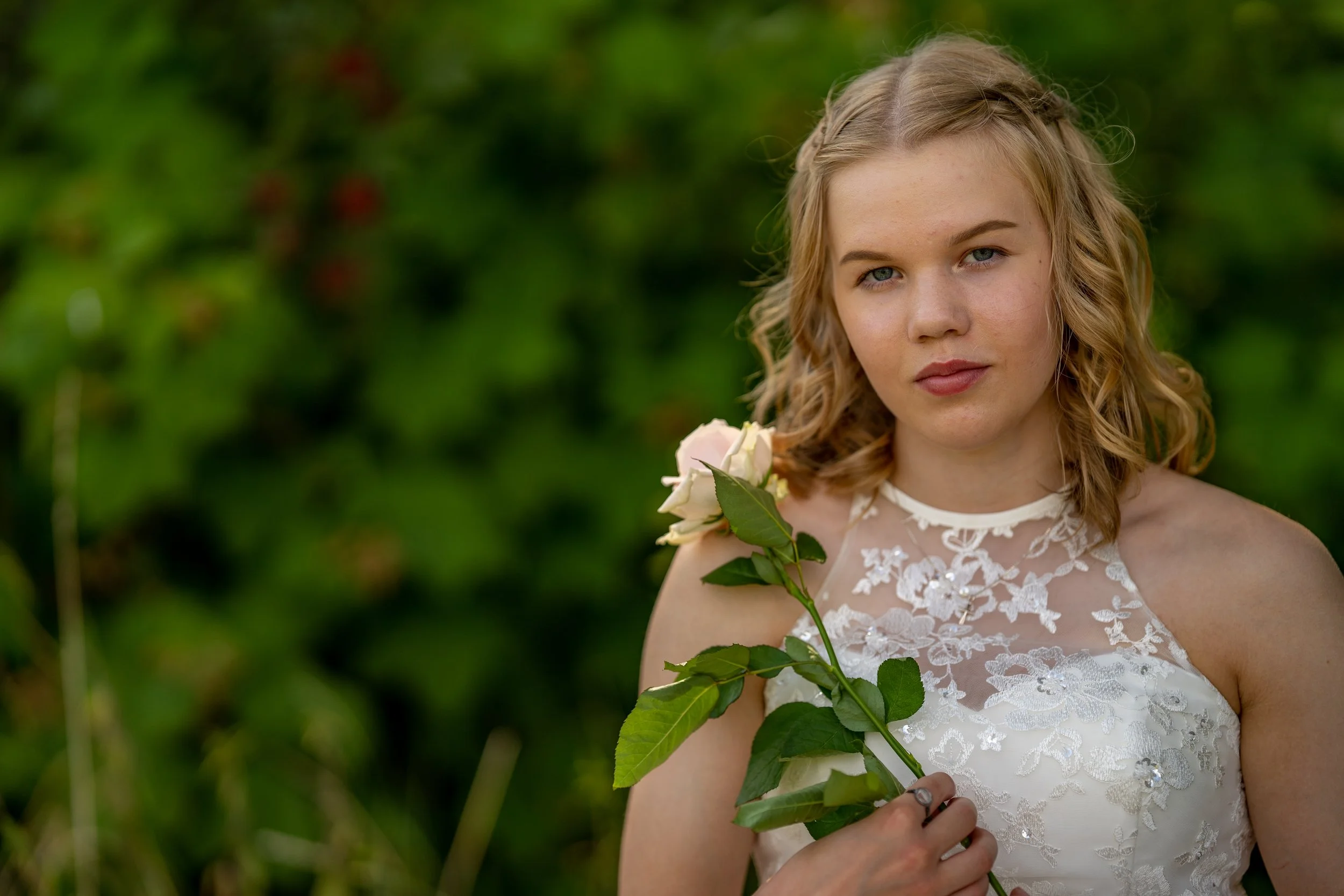 A young woman with blonde curly hair wearing a white lace dress, holding a pink rose, standing outdoors with green foliage in the background.