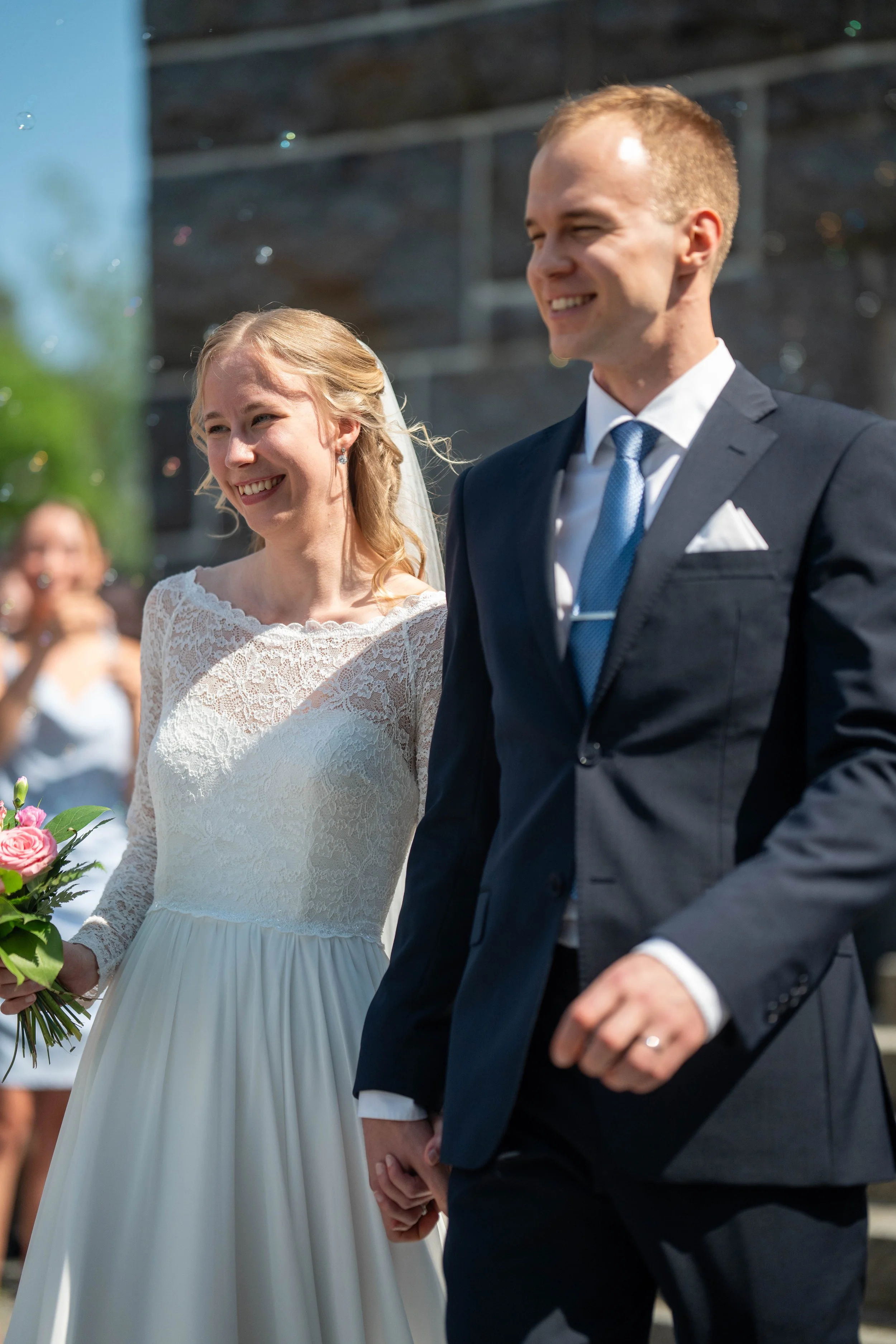 A smiling bride and groom holding hands during their wedding ceremony outdoors, with the bride holding a bouquet of pink roses.