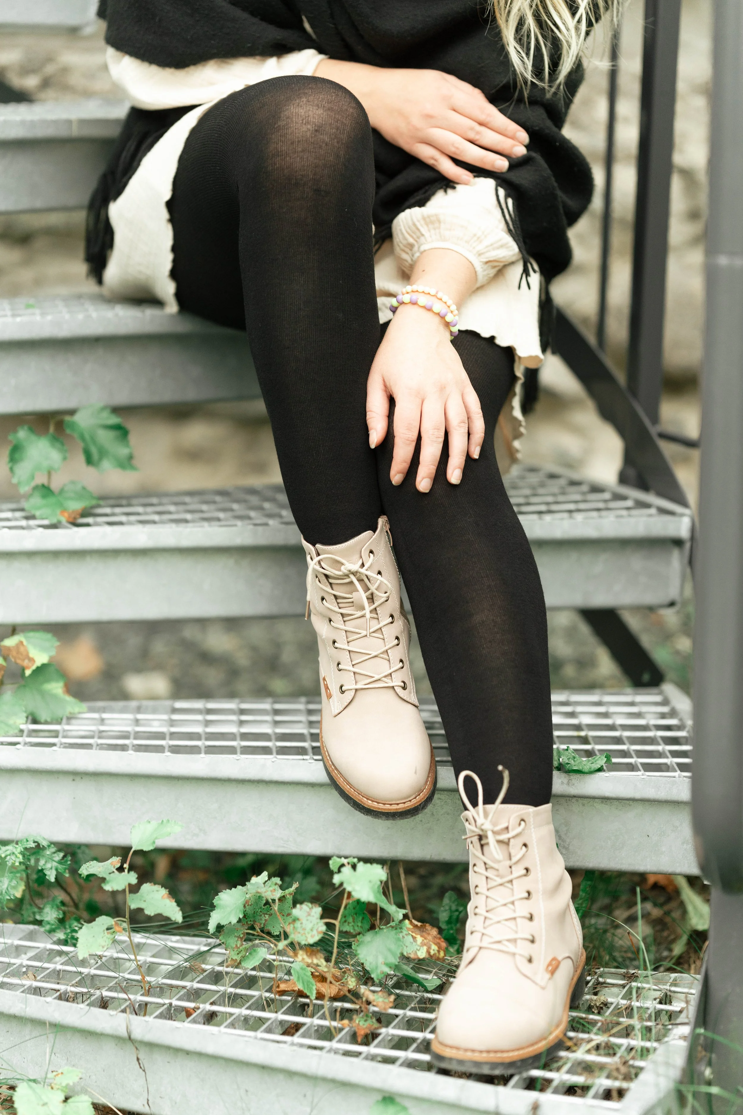 A person sitting on outdoor metal stairs, wearing black tights, beige boots, and a black and cream-colored outfit. Their face is not visible, and their right hand rests on their thigh, adorned with colorful beaded bracelets.