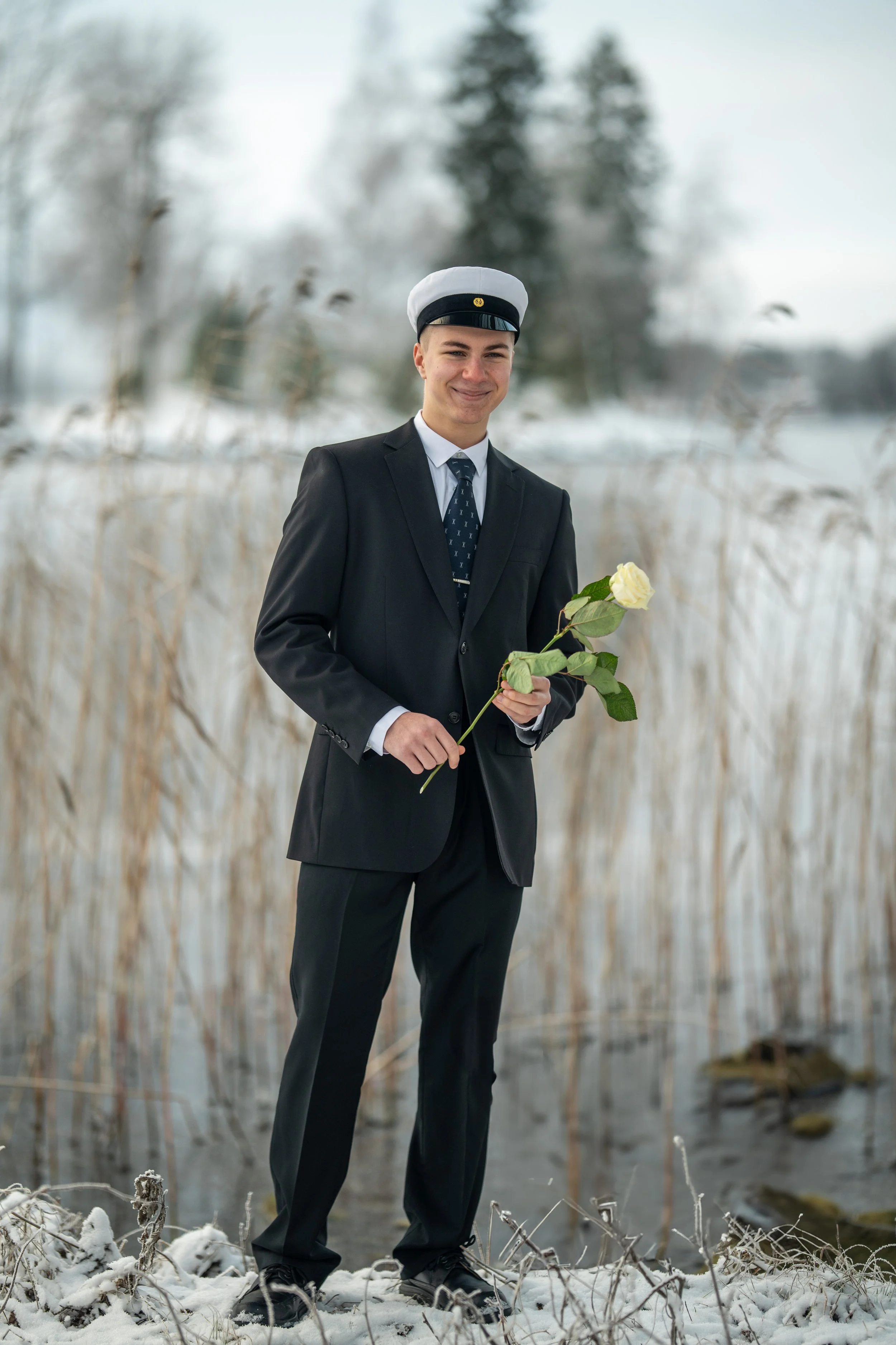 A young man dressed in a formal suit with a white graduation cap holding a white rose, standing outdoors in a snowy landscape near a body of water and tall grass, smiling.