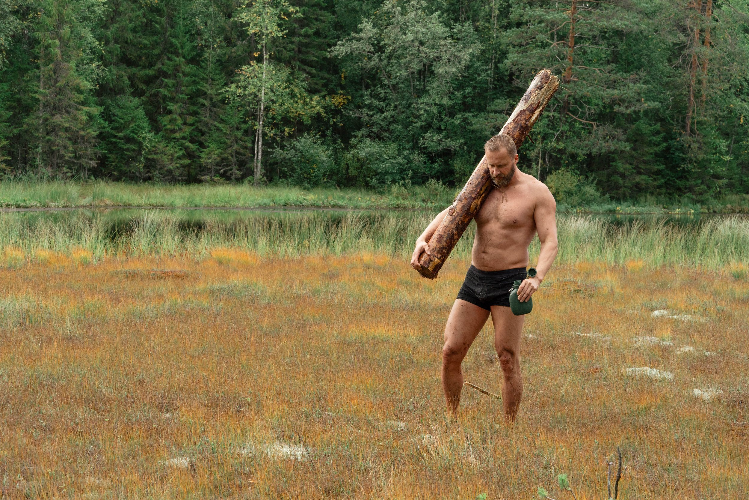 A man with a beard, shirtless, wearing black shorts, carrying a large log on his right shoulder and holding a green object in his left hand, standing in a grassy wetland area with trees in the background.