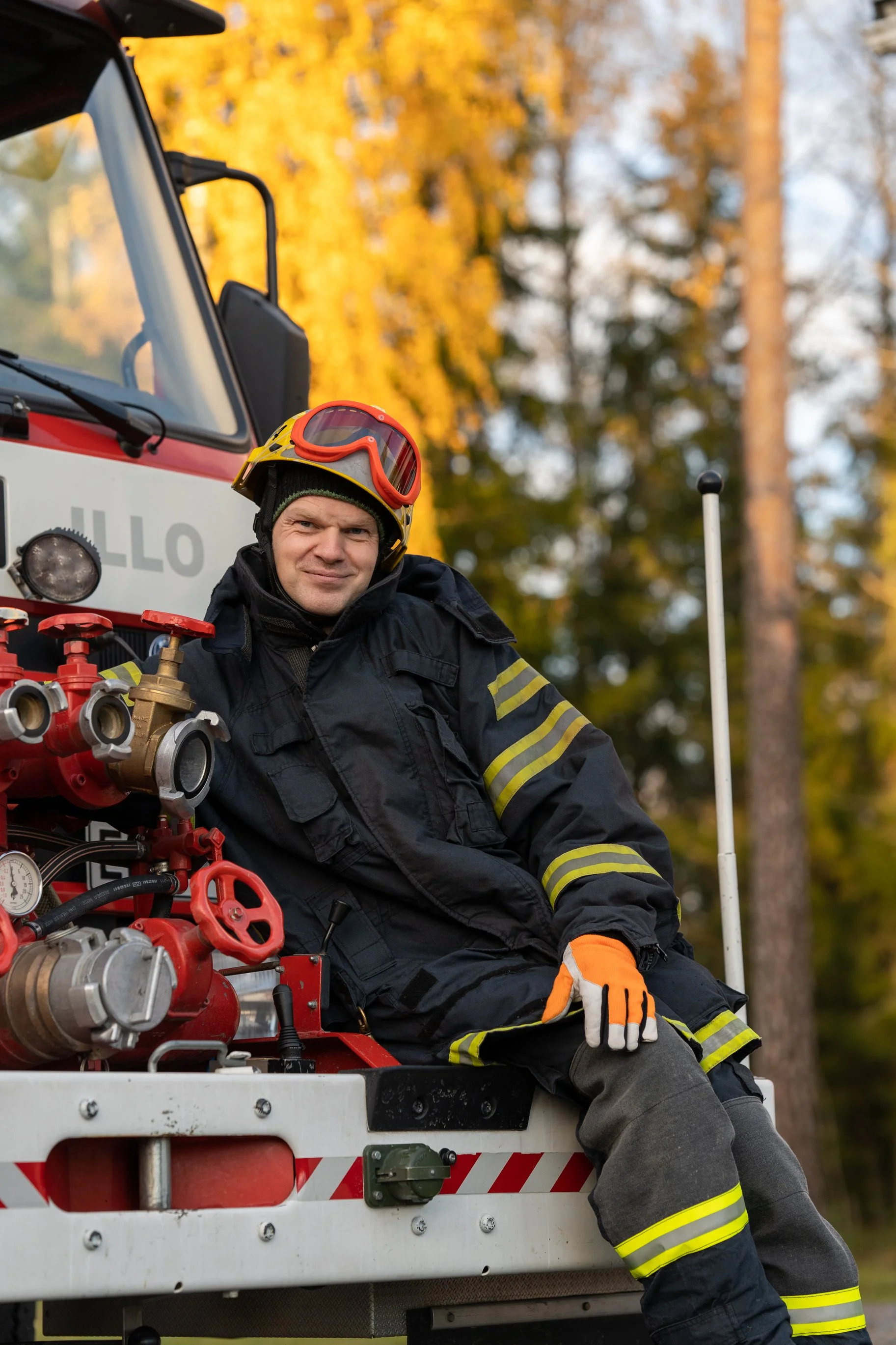 Firefighter sitting on a fire truck outdoors during autumn, wearing protective gear and an orange glove, with trees and yellow leaves in the background.