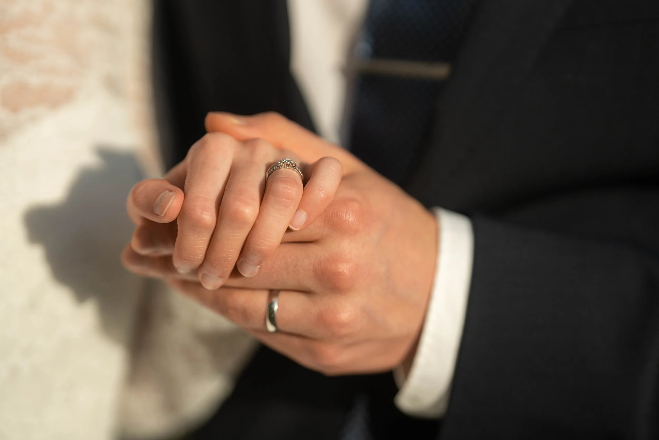 Close-up of a person's hands clasped together, wearing wedding rings and a dark suit with a white shirt.