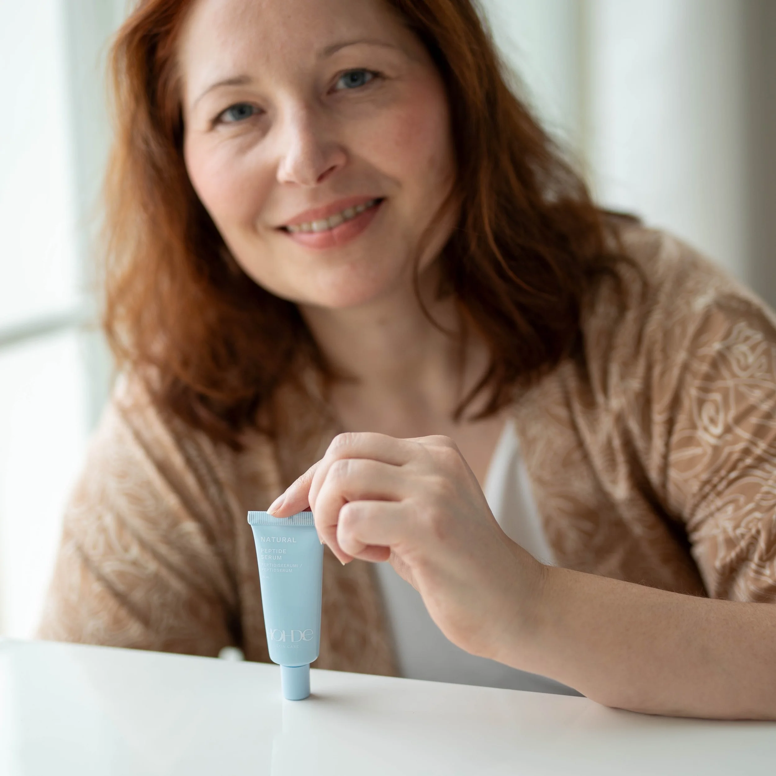 A woman with red hair is smiling and holding a small blue tube of skincare product on a white surface, near a window with natural light.