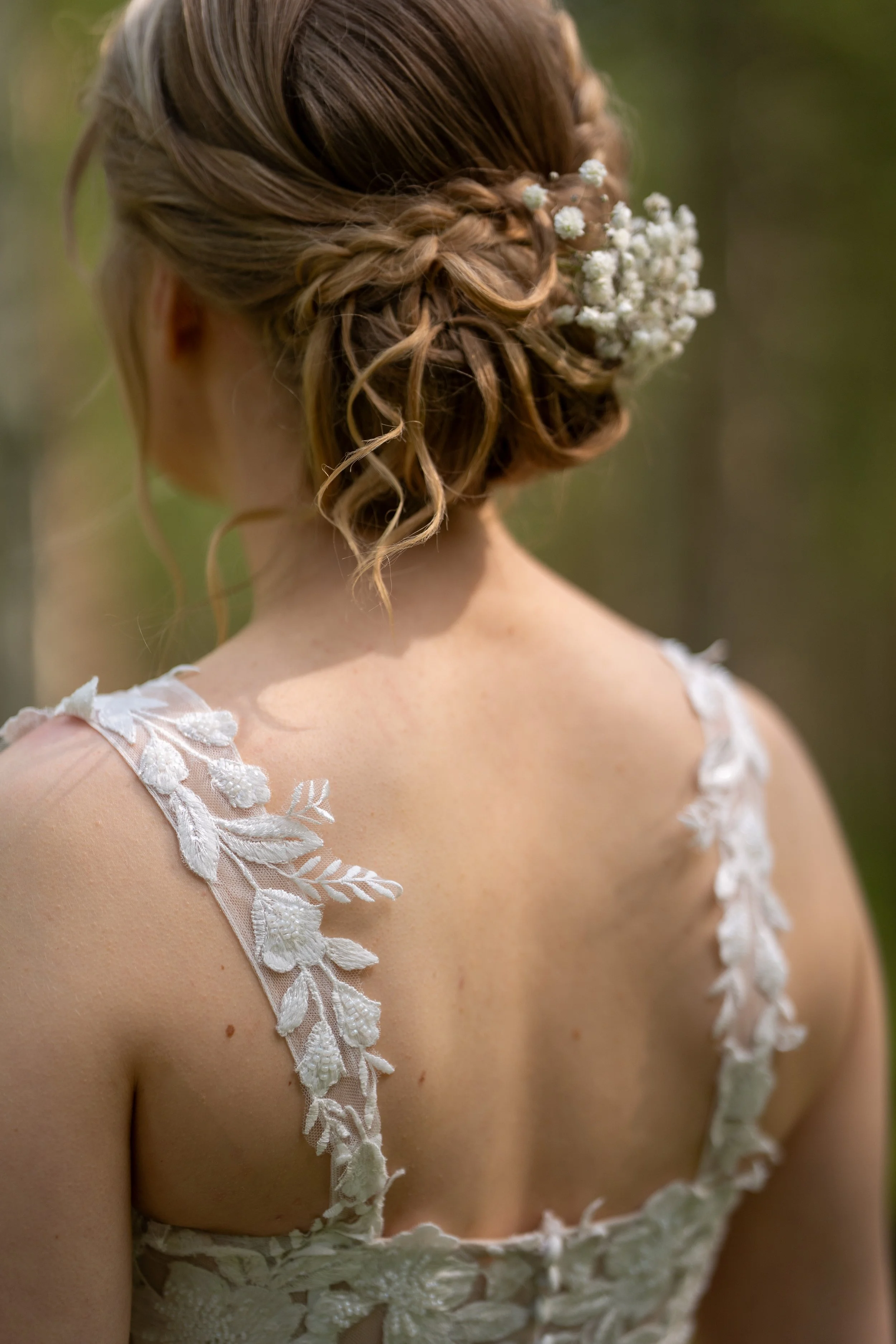 A woman with curly, styled hair decorated with white flowers, wearing a white lace dress with embroidered floral details on the shoulder straps, standing outdoors.