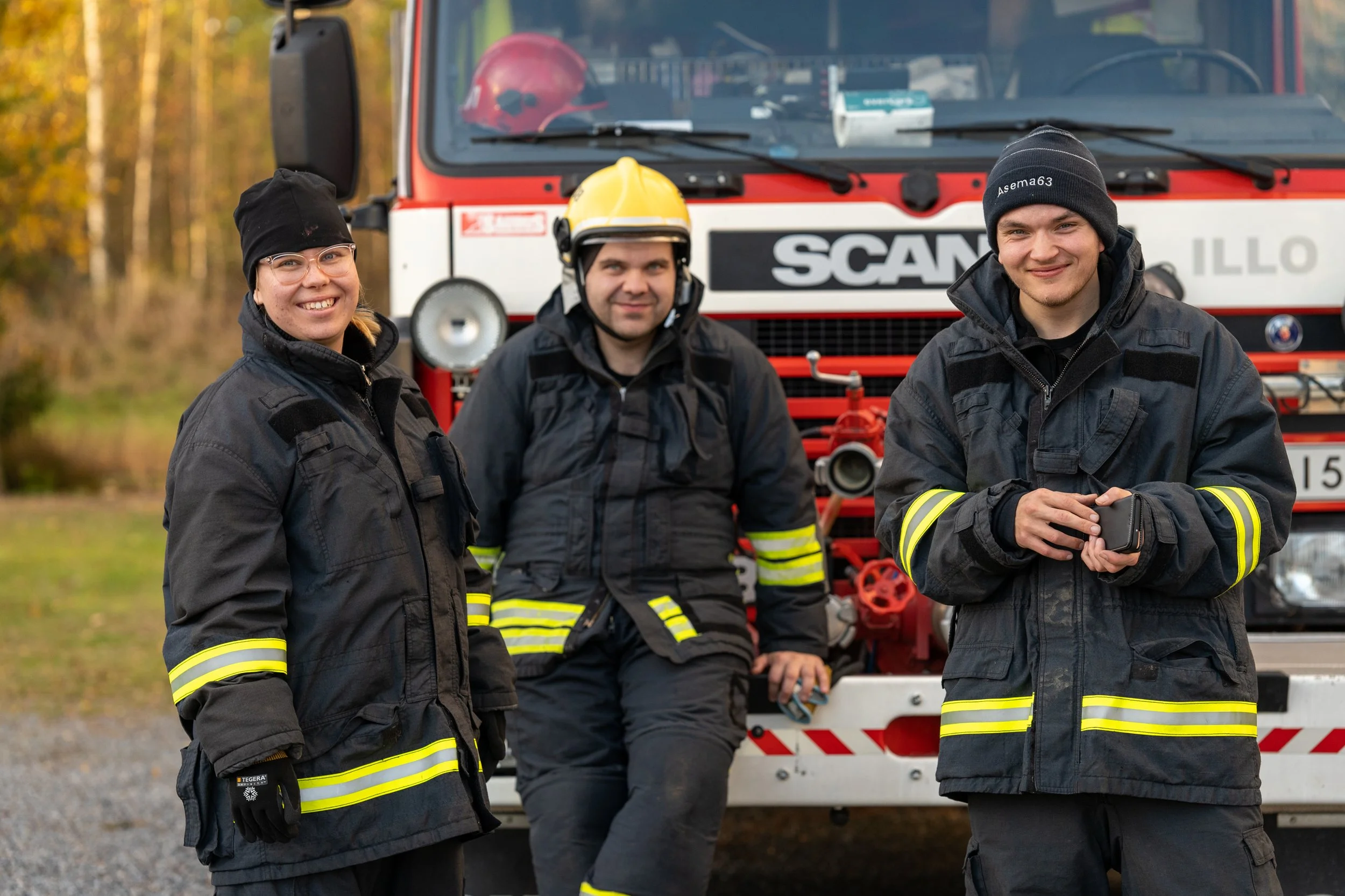 Three firefighters standing outdoors in front of a fire truck. They are dressed in black firefighting gear with reflective yellow stripes. One firefighter, wearing a helmet, is leaning against the truck, while the other two are smiling and holding a 
