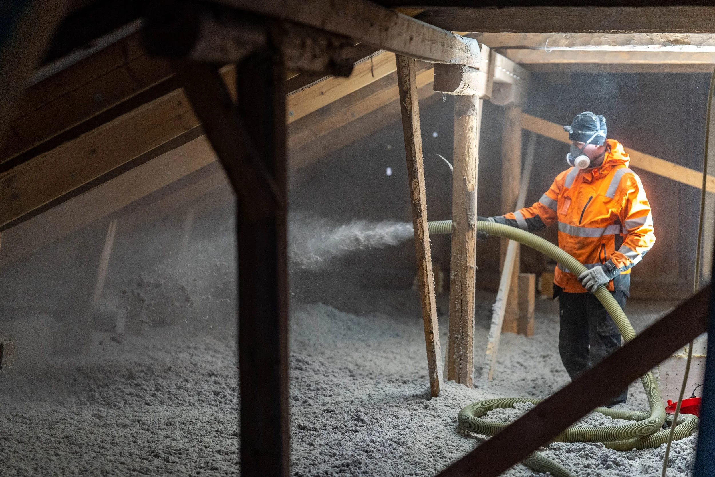 Worker in an orange safety jacket and protective mask operating a hose to spray a granular substance inside an attic or construction space.