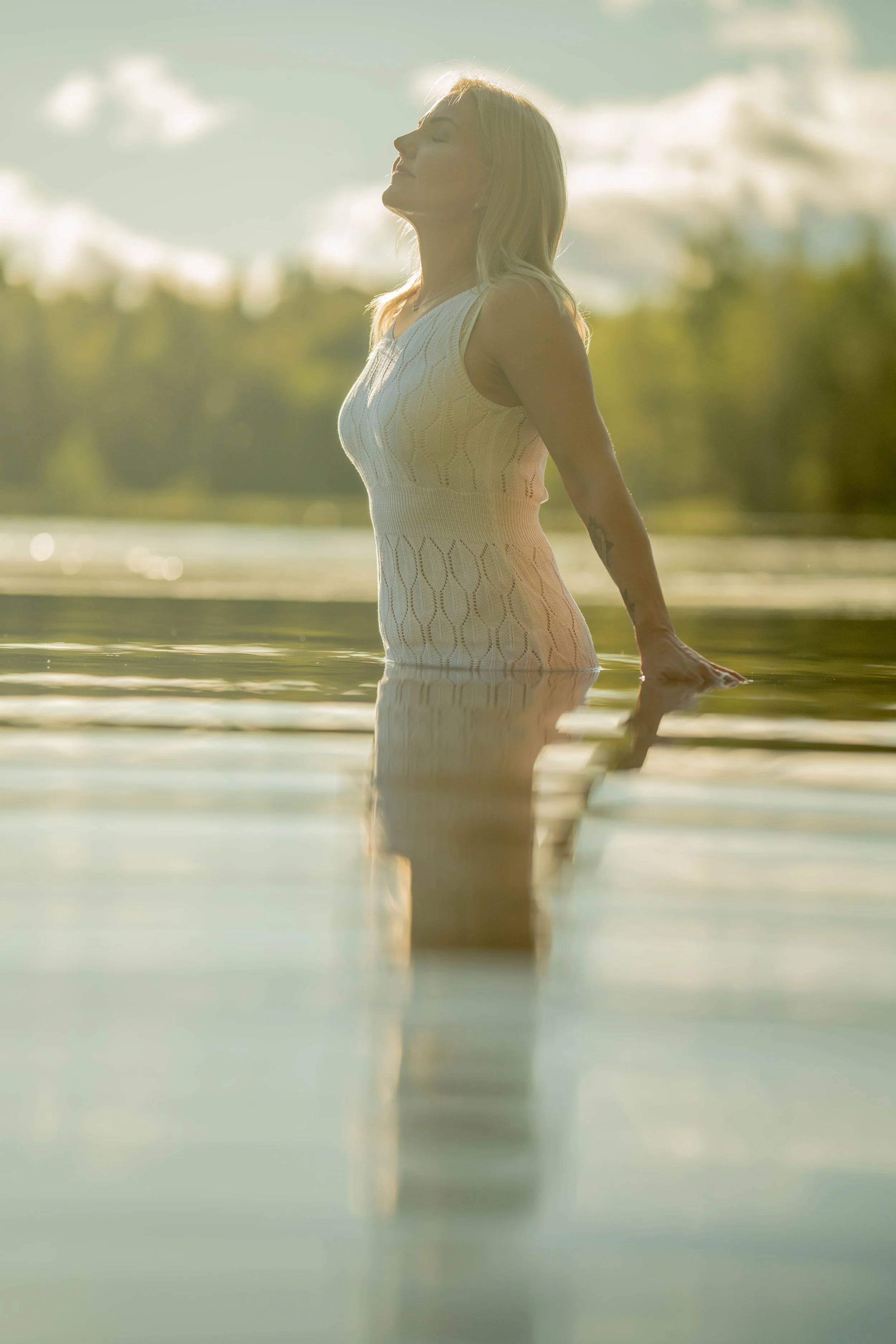 A woman standing in a body of water, enjoying the outdoors with her eyes closed, wearing a sleeveless top, during sunset or sunrise.
