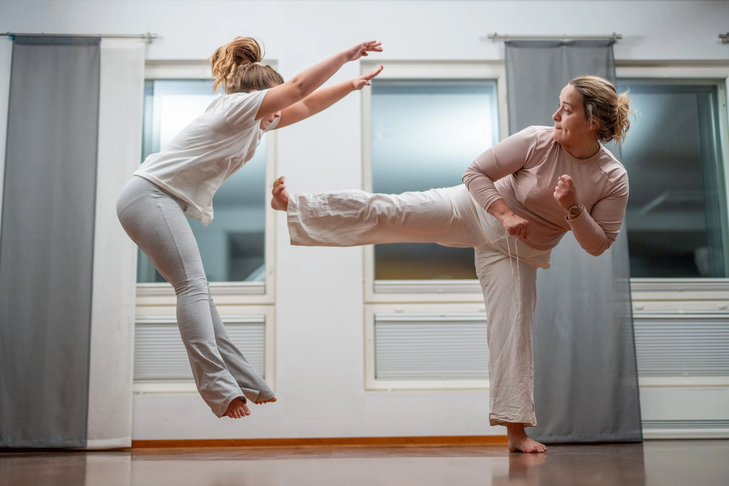 Two women practicing martial arts in a room with large windows, one woman kicking while the other blocks.