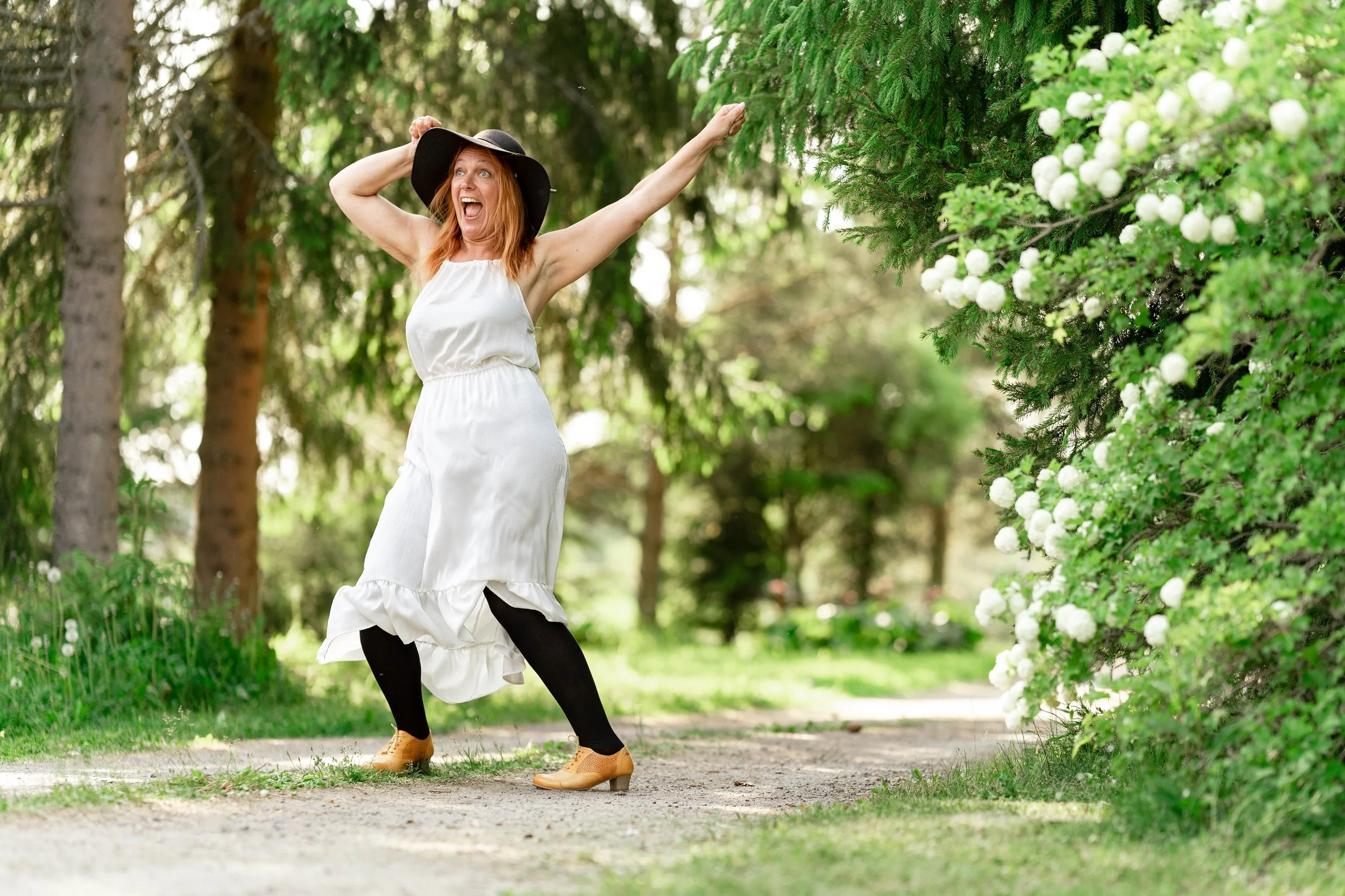A woman with red hair, wearing a black sunhat, white dress, black leggings, and tan shoes, stands in a cheerful pose on a dirt path in a lush green park with trees and white flowering bushes.