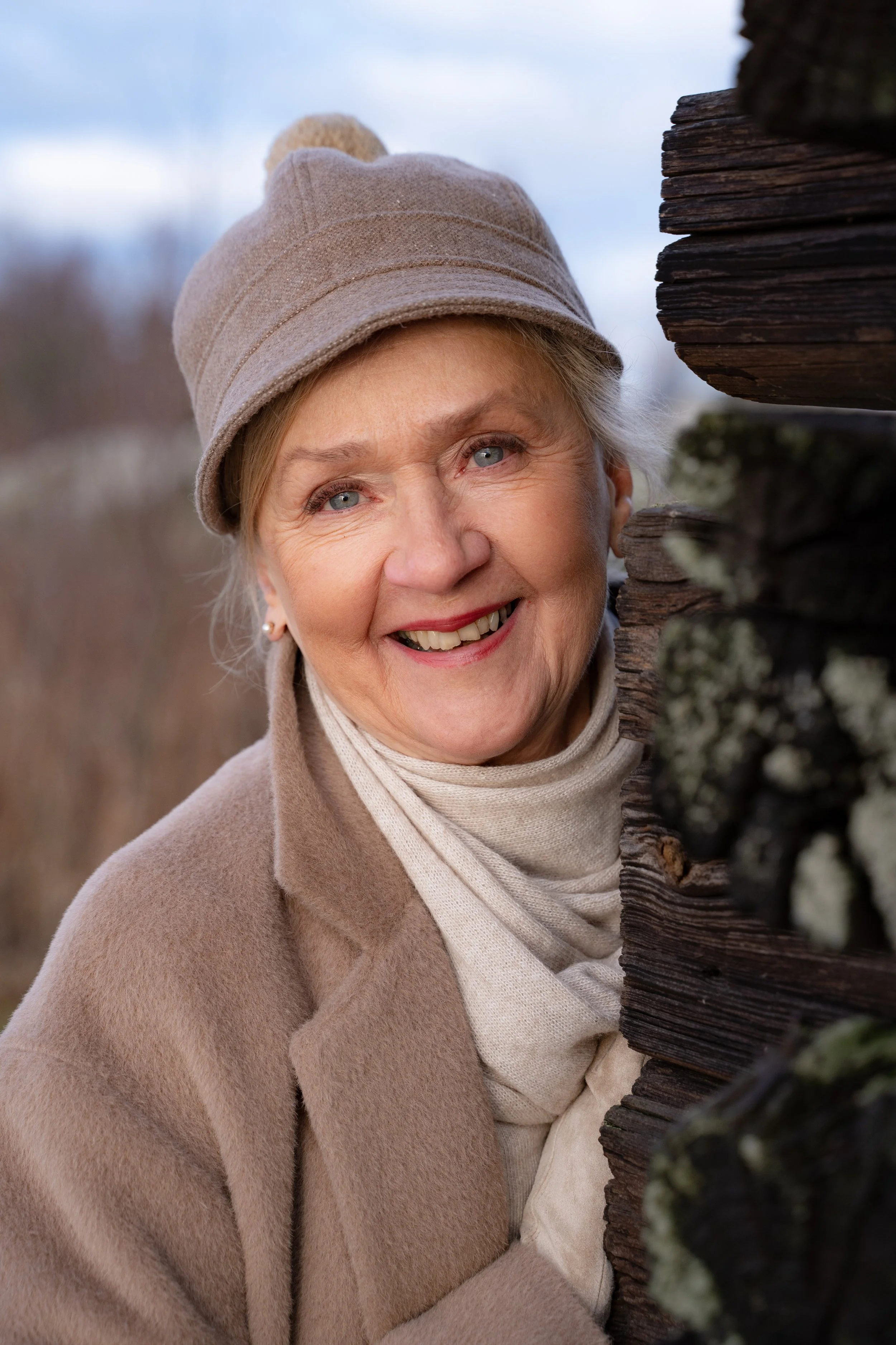 A smiling elderly woman in a beige coat, scarf, and knit cap standing outdoors near a wooden structure, with blurred trees and sky in the background.