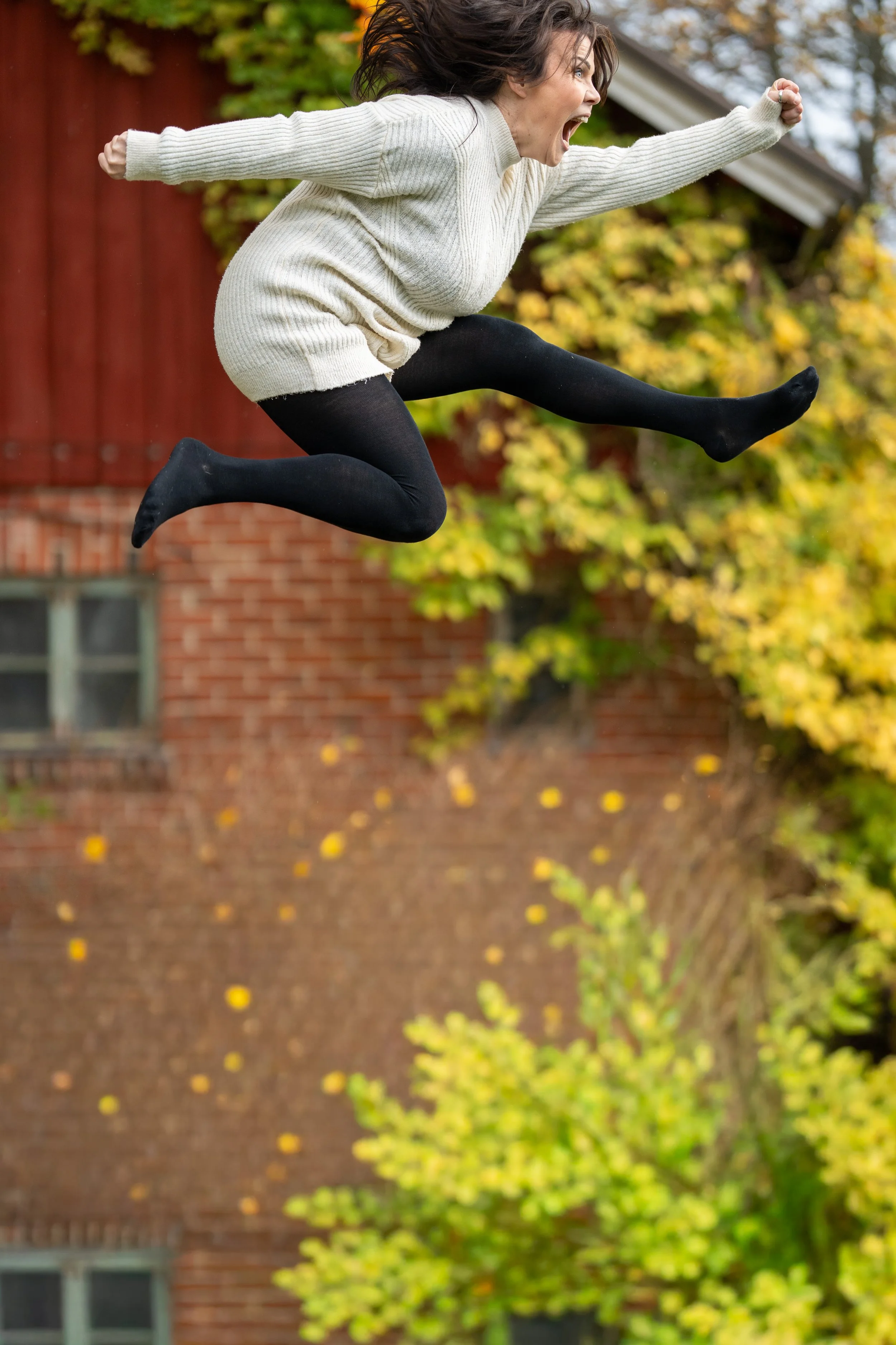 A woman in a cream sweater and black tights jumps in the air with her arms outstretched and mouth open, outdoors with autumn trees and a red brick building in the background.