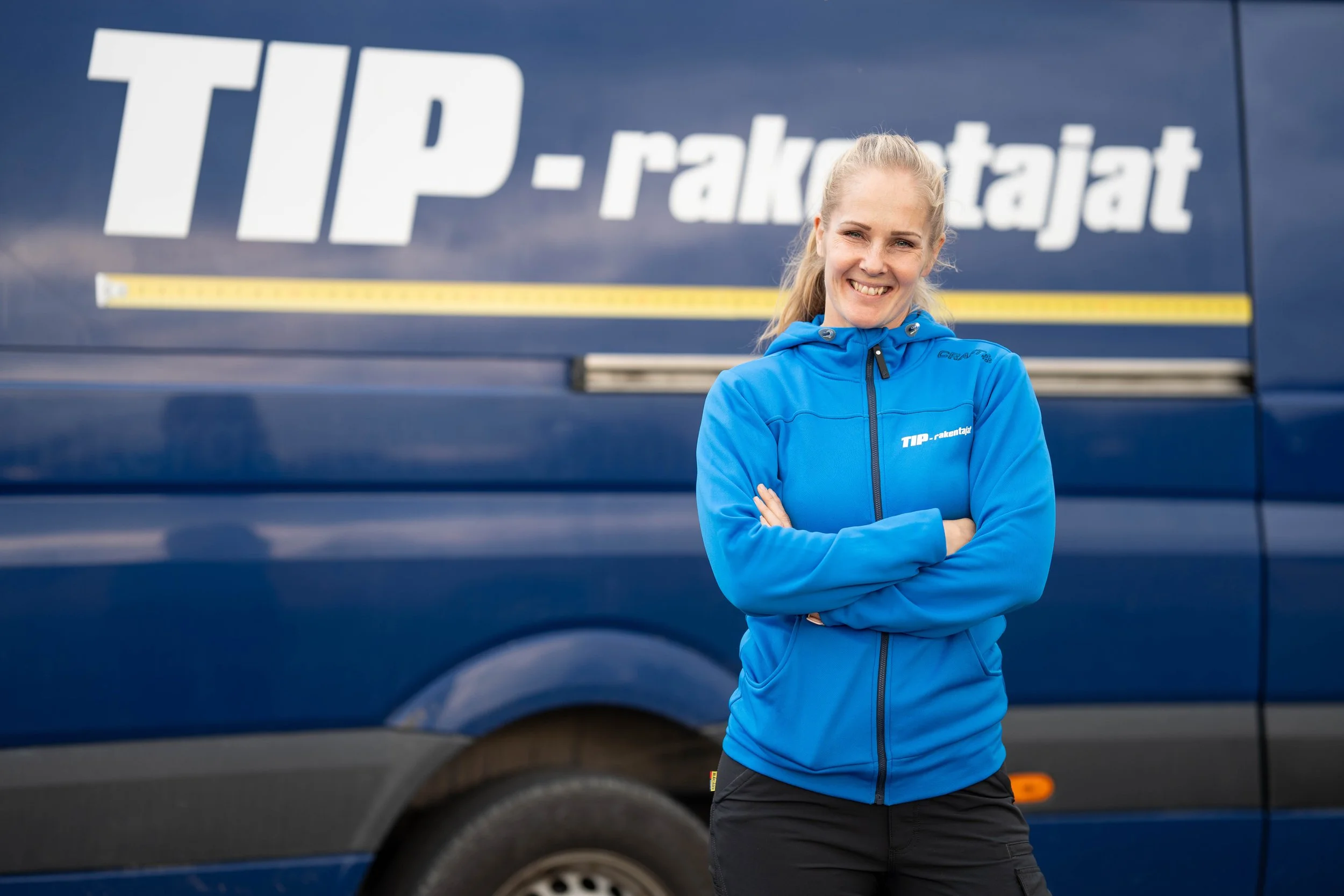 A smiling woman in a blue jacket standing with arms crossed in front of a blue van marked "TIP-rakentajat".