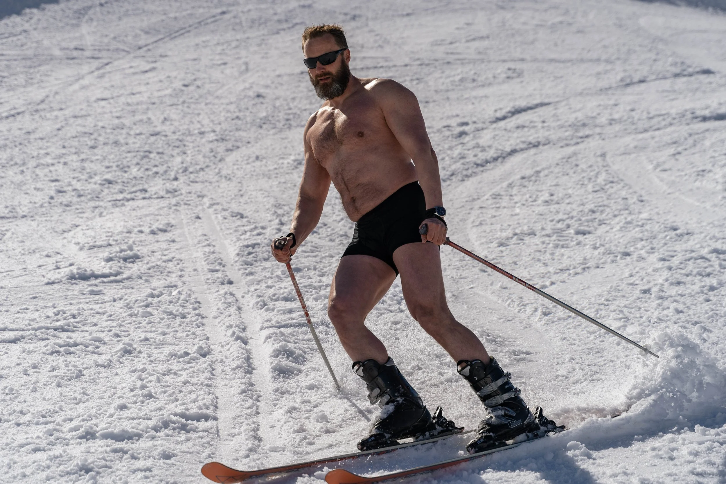 Man skiing on snow, shirtless, wearing black shorts, sunglasses, and a watch, holding ski poles.