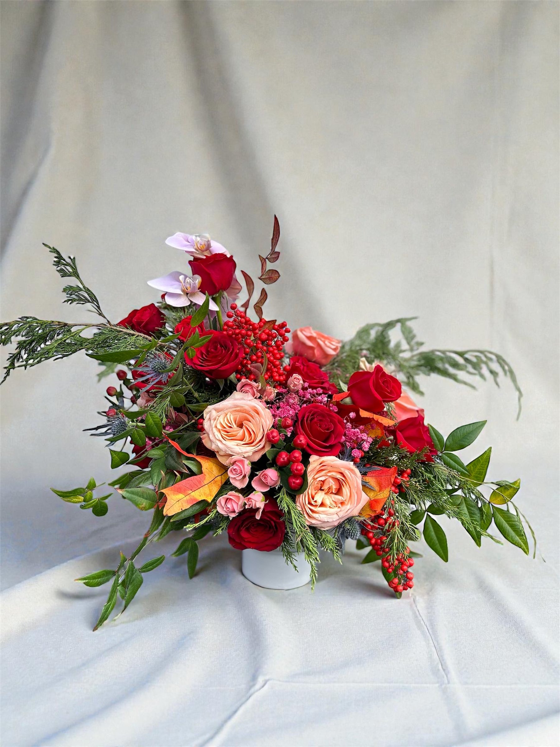 A colorful flower arrangement with pink, red, and peach roses, purple orchids, red berries, green leaves, and assorted greenery in a white vase.
