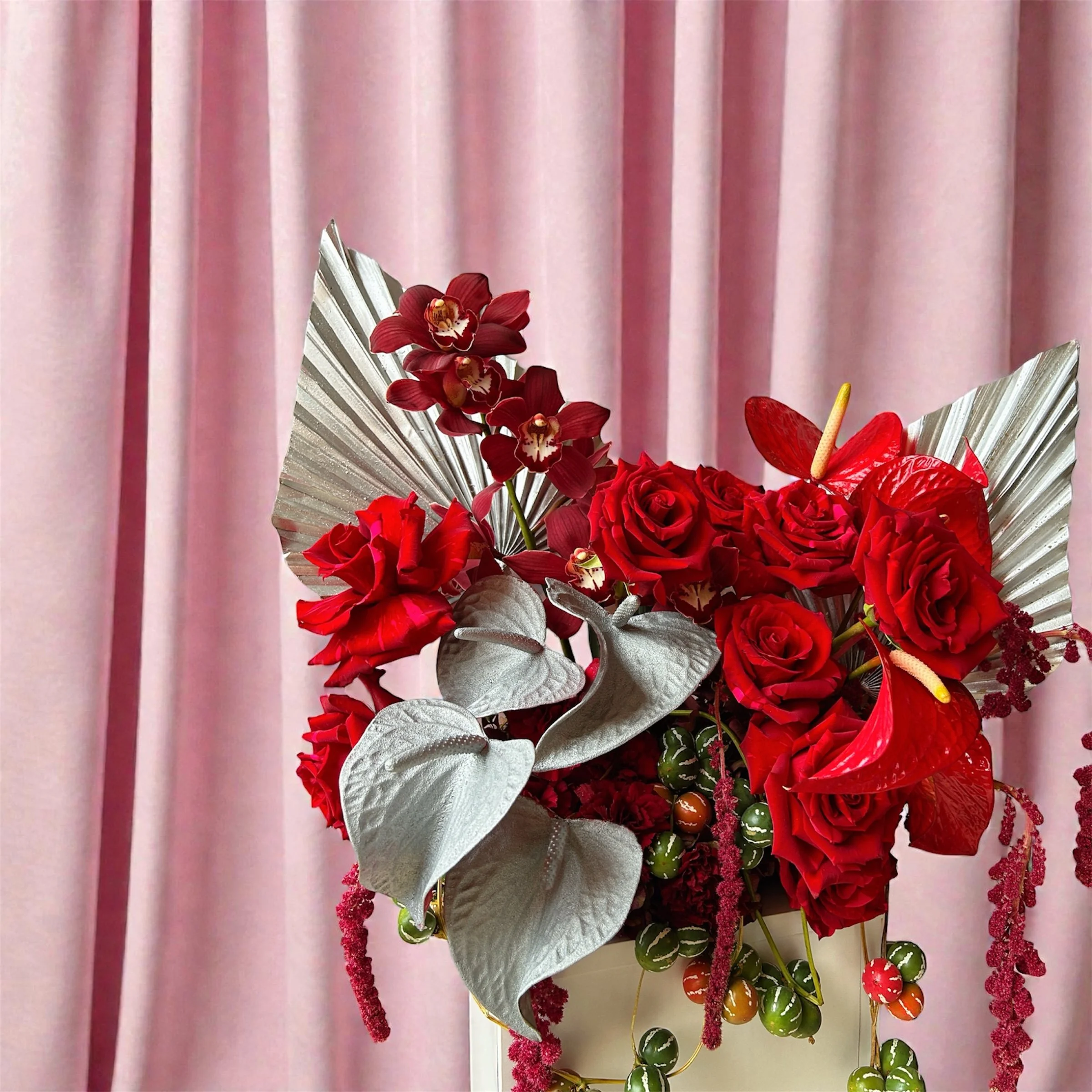 A floral arrangement with red roses, red orchids, red anthuriums, and silver leaves against a pink curtain background.