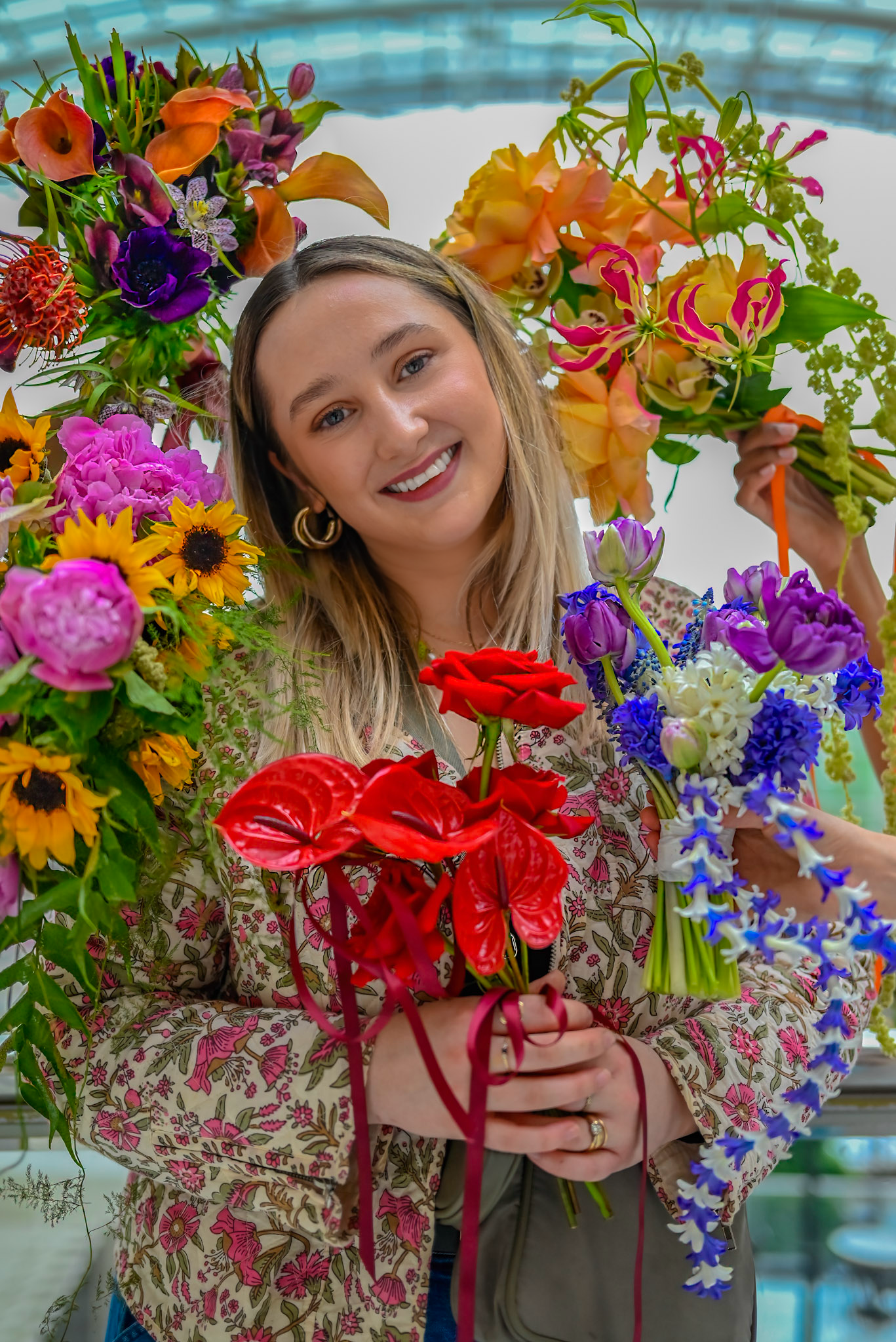 A woman smiling and holding colorful flower bouquets surrounded by vibrant flowers including roses, lilies, and other blooms.