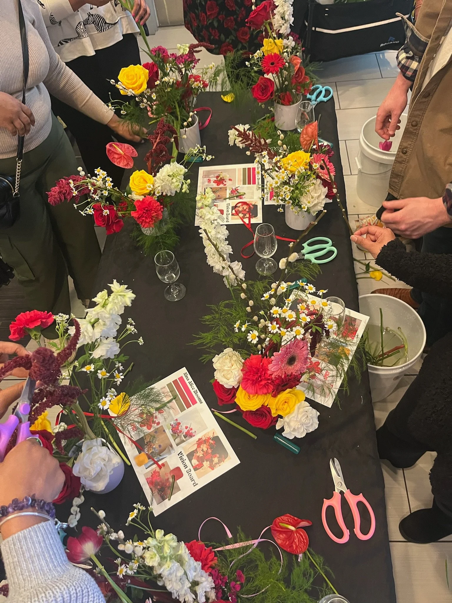 People gathered around a table creating flower arrangements with various colorful flowers, scissors, and instructional sheets.