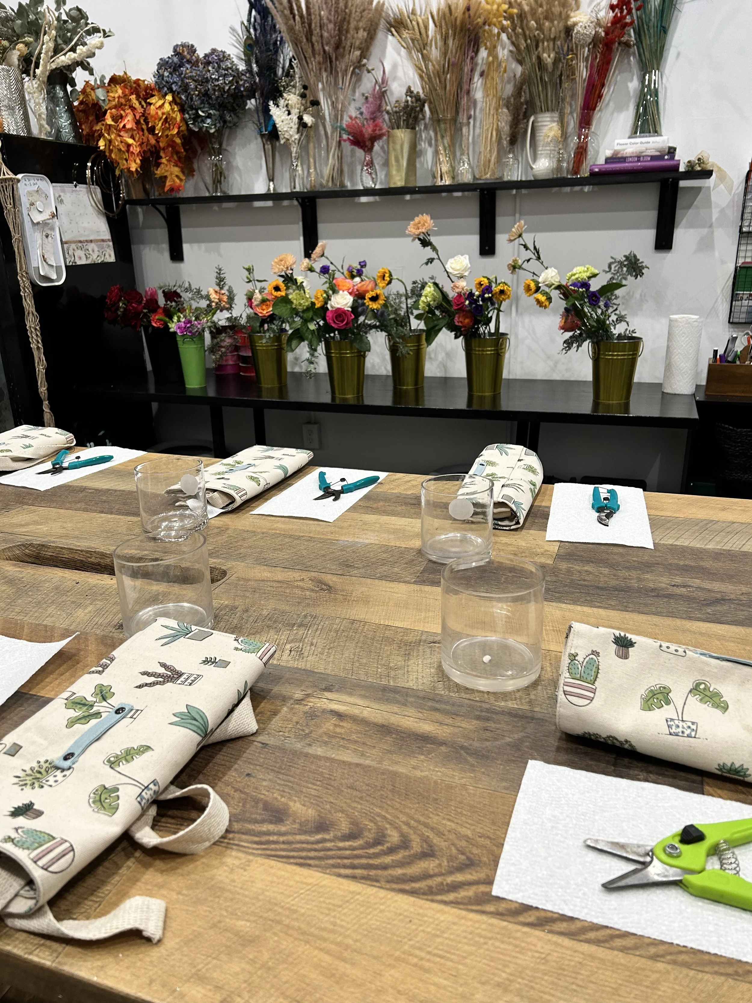 A workshop or craft room with a wooden table set for a floral arranging activity, featuring rolled towels with cactus patterns, clear glasses, and green-handled scissors. In the background, there are colorful bouquet arrangements in green vases, drie