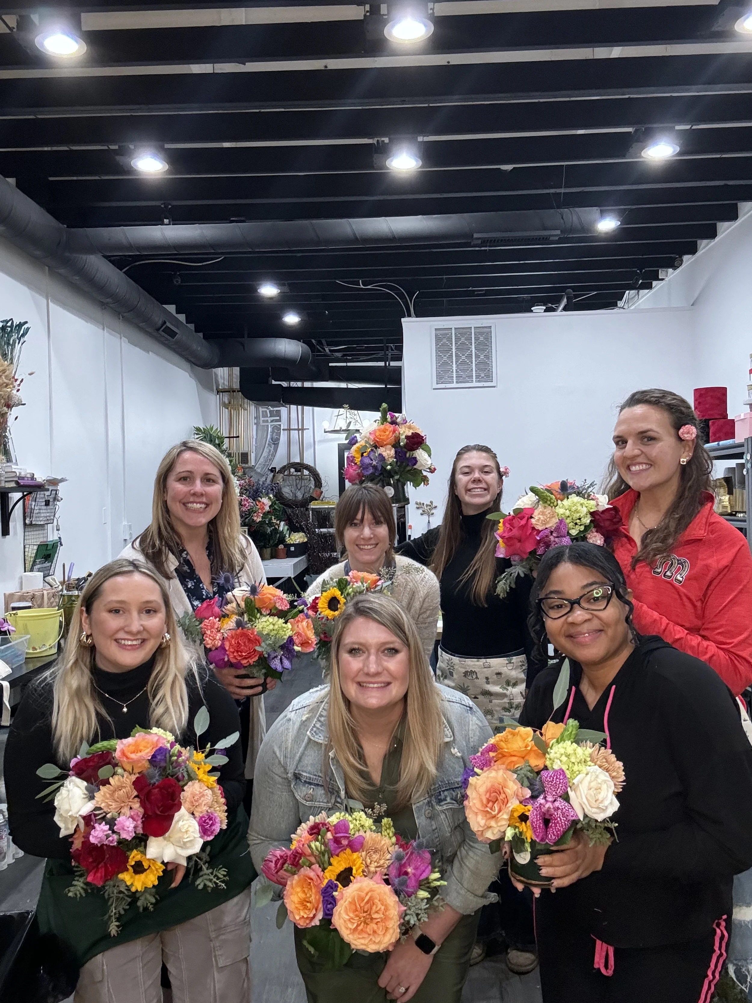 Group of seven women holding colorful flower bouquets in a floral shop, smiling at the camera.