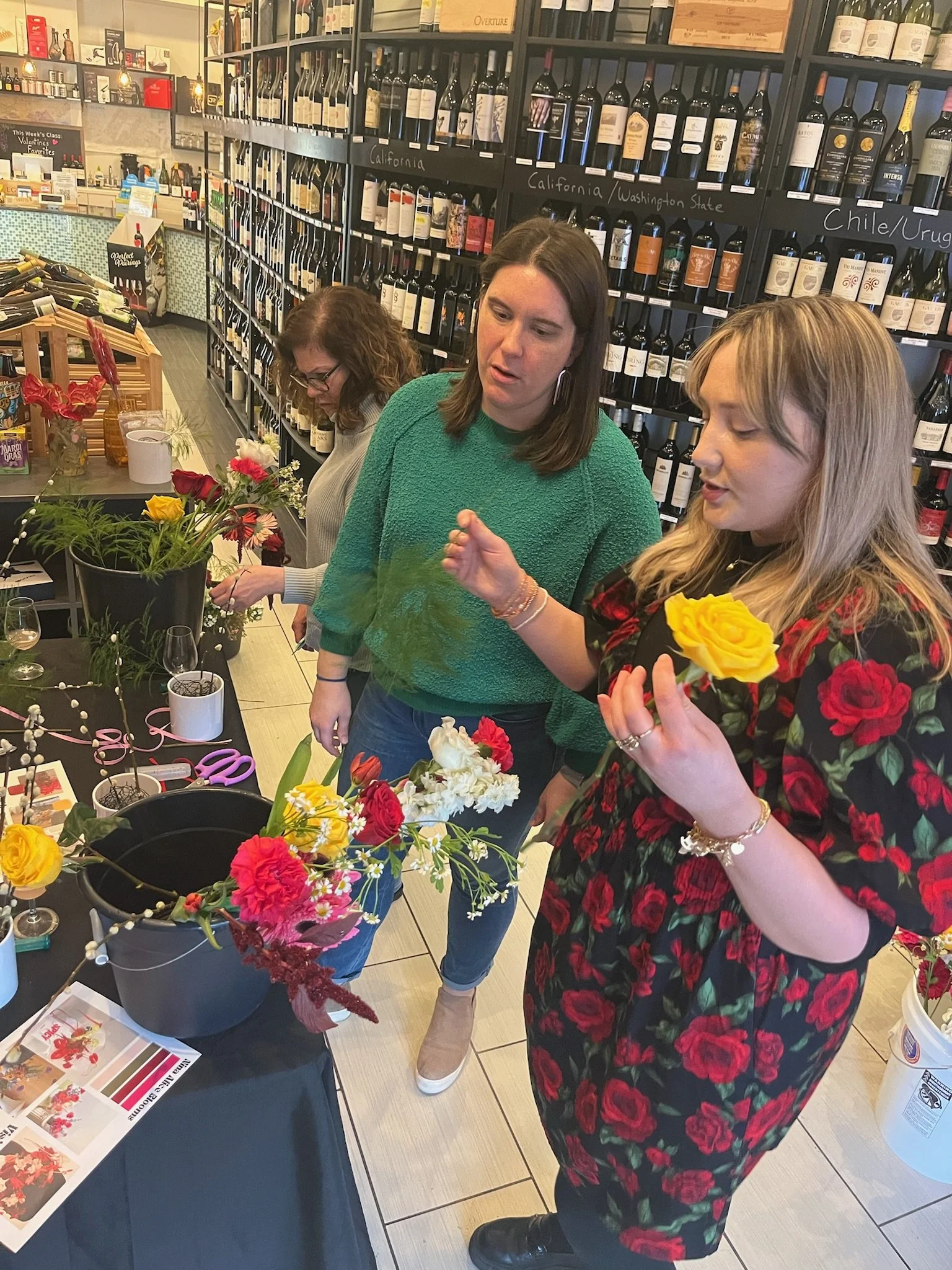 Three women are arranging flower bouquets at a table inside a wine shop, with shelves of wine bottles behind them.