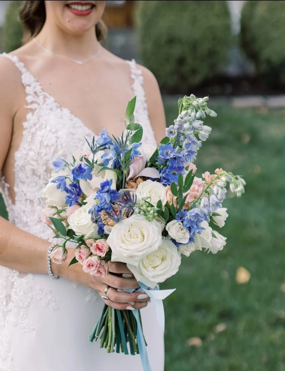 A woman in a wedding dress holding a bouquet of white, pink, and blue flowers outdoors.