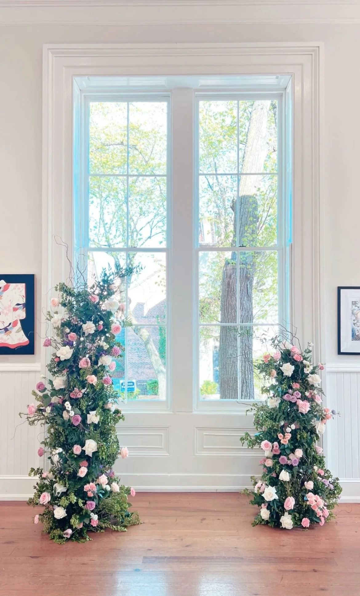Two floral arrangements with pink and white roses positioned on either side of a large white-framed window with a view of trees outside.
