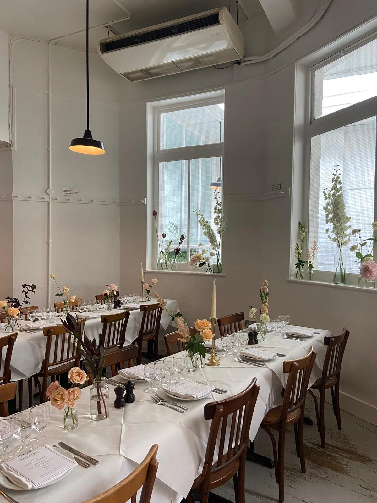 A decorated dining room with long tables covered in white tablecloths, set with glassware, plates, and silverware. Flower vases and tall candles are arranged along the tables. The room has large windows with flower arrangements inside, white walls, and wooden chairs around the tables. Overhead lighting and an air conditioning unit are also visible.