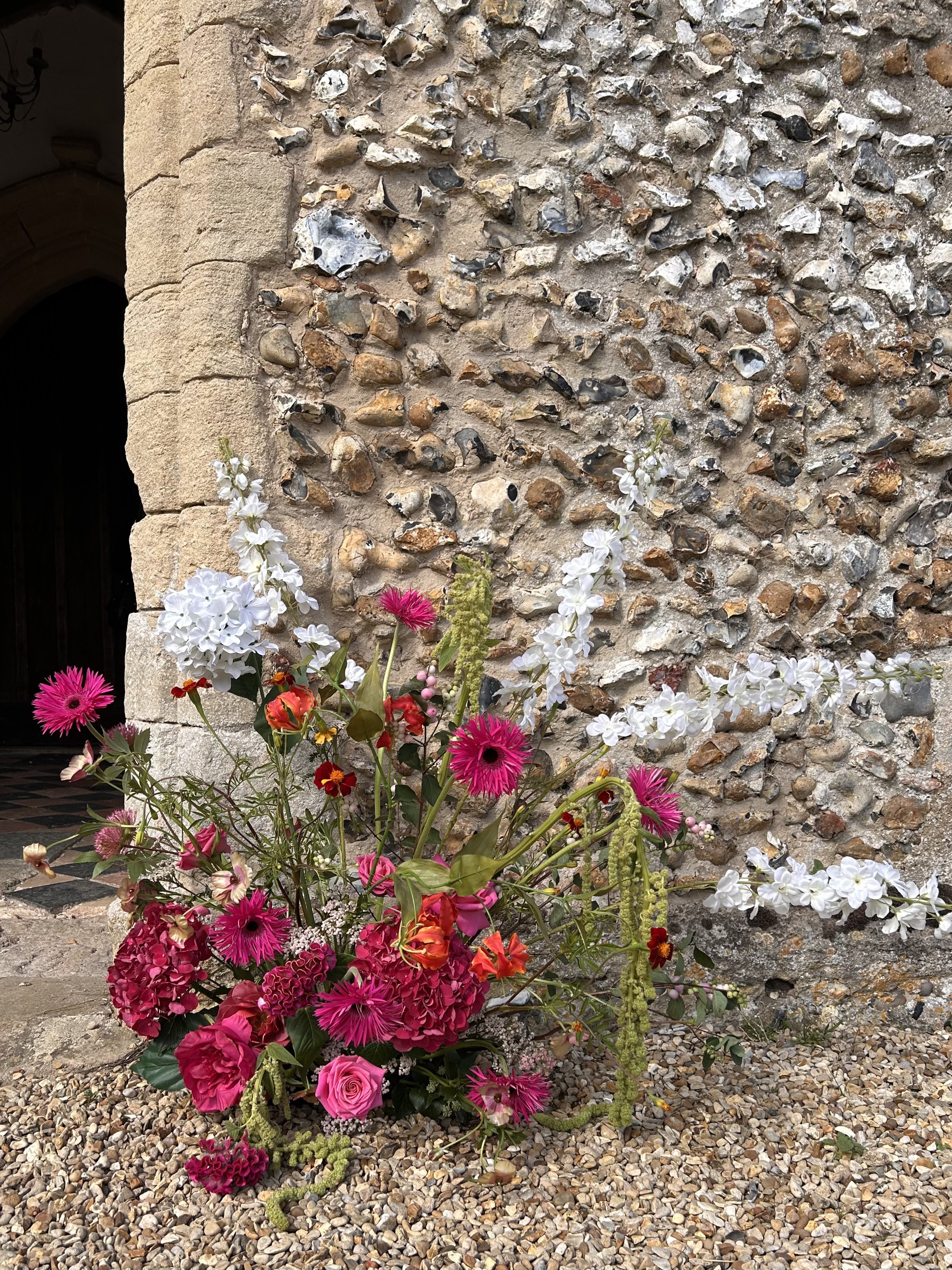 Colorful bouquet of pink, white, and red flowers placed on gravel ground against a stone wall of a building with an arched doorway.