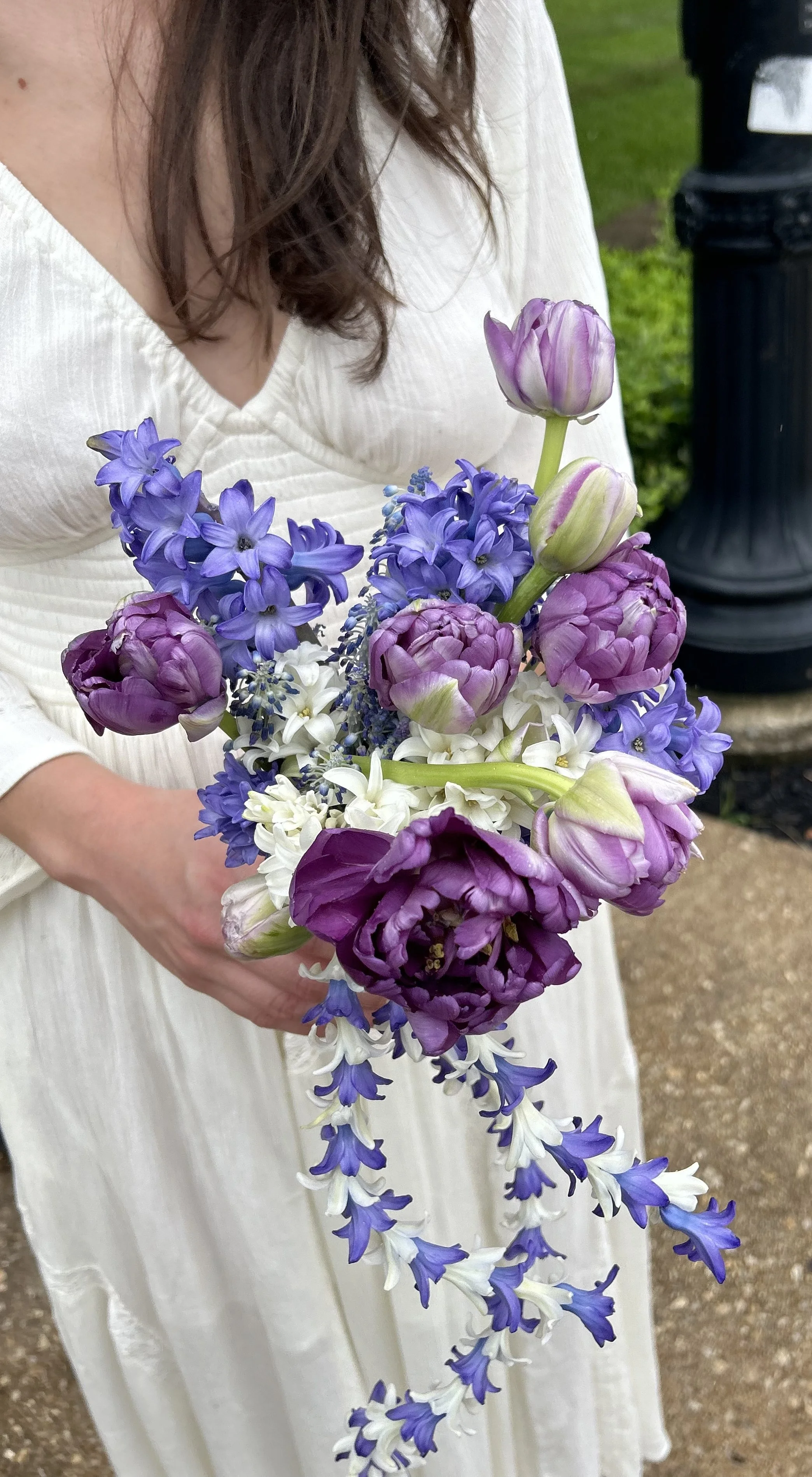 A person holding a bouquet of purple, white, and pink flowers, including hyacinths, tulips, and possibly bluebells, with the person wearing a white dress.