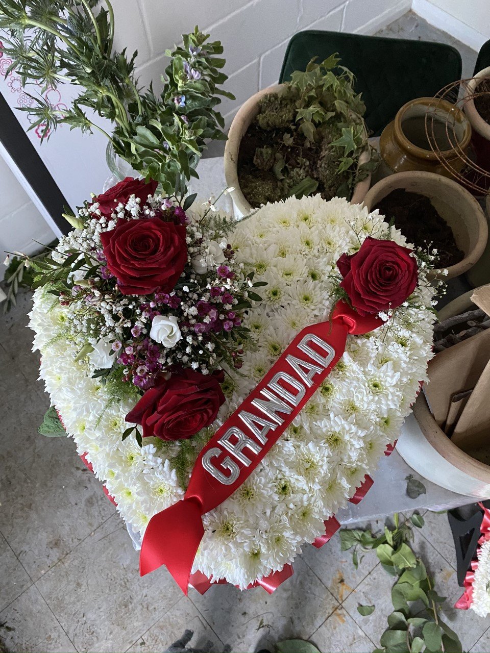 A heart-shaped floral arrangement with white flowers, decorated with three red roses, small purple and white flowers, and greenery. It has a red ribbon with