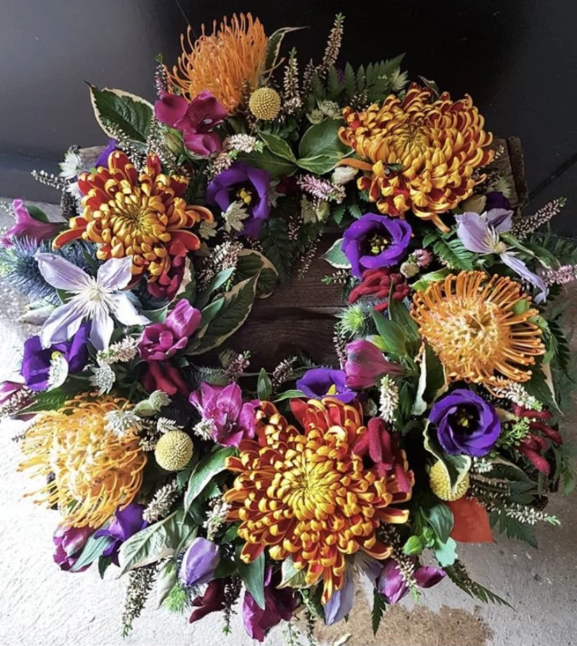 Colorful flower arrangement with orange, purple, pink, and white blooms, including chrysanthemums, lilies, and other mixed flowers, set against a dark background.