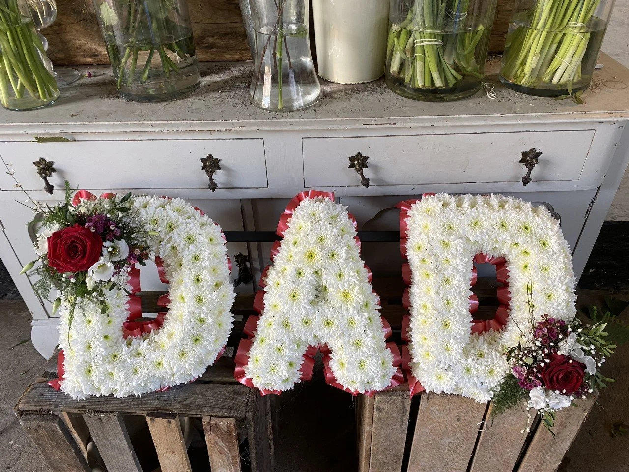 Floral arrangement spelling out the word 'LOVE' in white flowers with red and white roses, placed on a wooden crate in front of a whiteFurniture cabinet with glass vases of flowers on top.