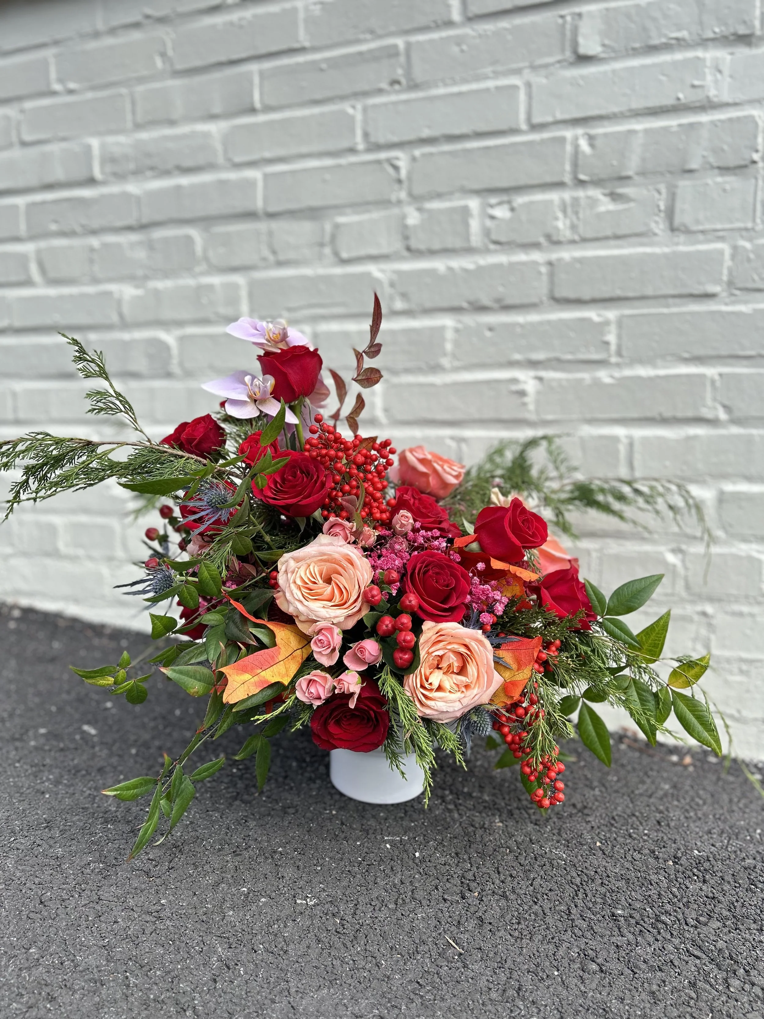 A floral arrangement with roses, orchids, berries, and greenery in a white vase on a dark asphalt surface against a white brick wall.