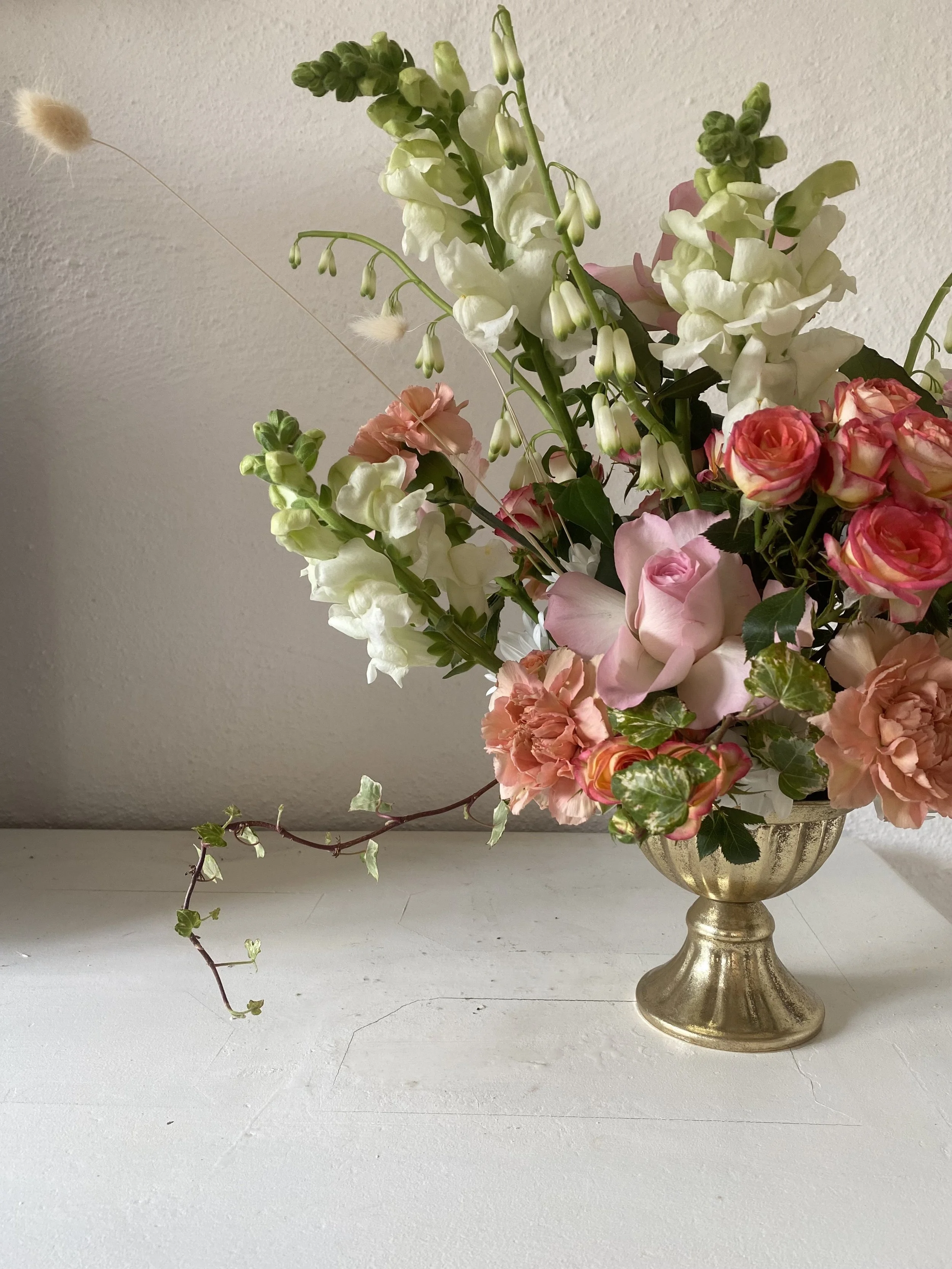 A floral arrangement in a gold vase featuring pink, white, and peach flowers with green leaves, set on a white surface against a plain wall.