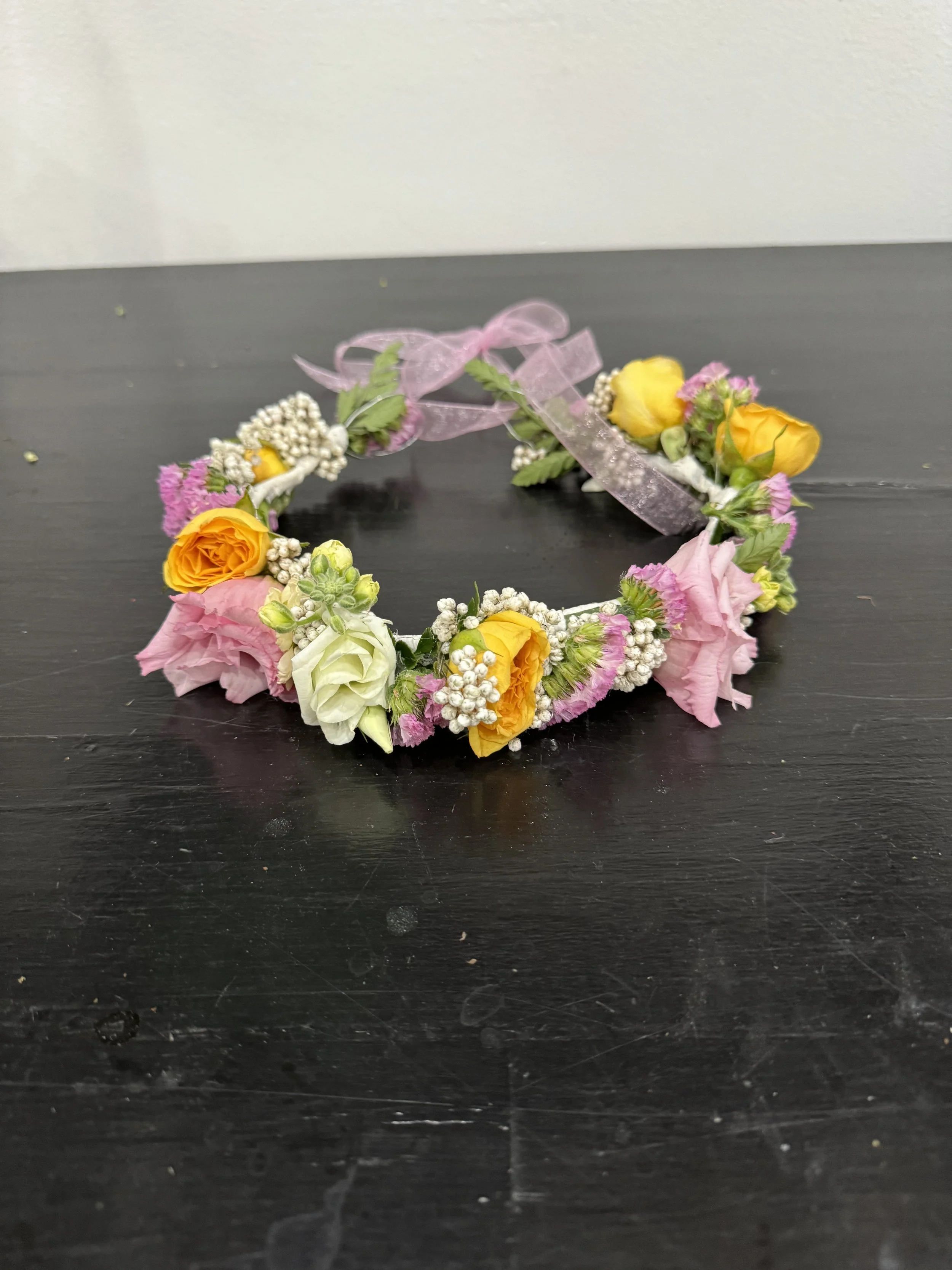 A flower crown made of pink, yellow, and white flowers with greenery, decorated with a pink ribbon.