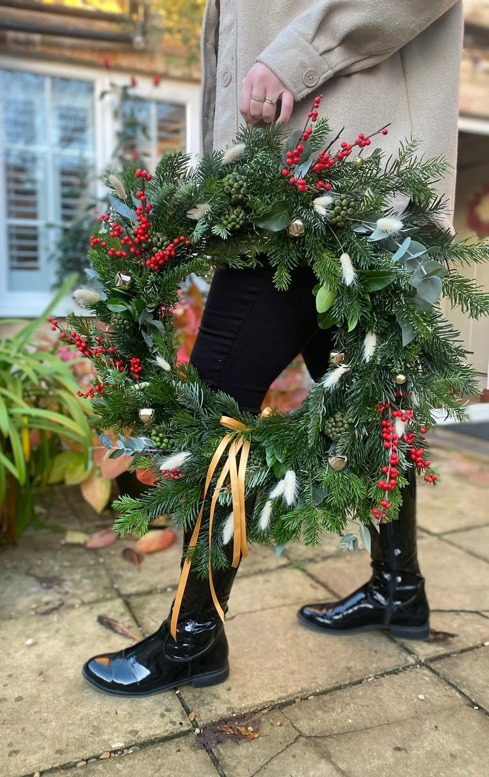 Person standing outdoors on a brick patio holding a large Christmas wreath decorated with evergreen branches, red berries, small bells, and white feathers, dressed in black boots, black pants, and a tan coat.